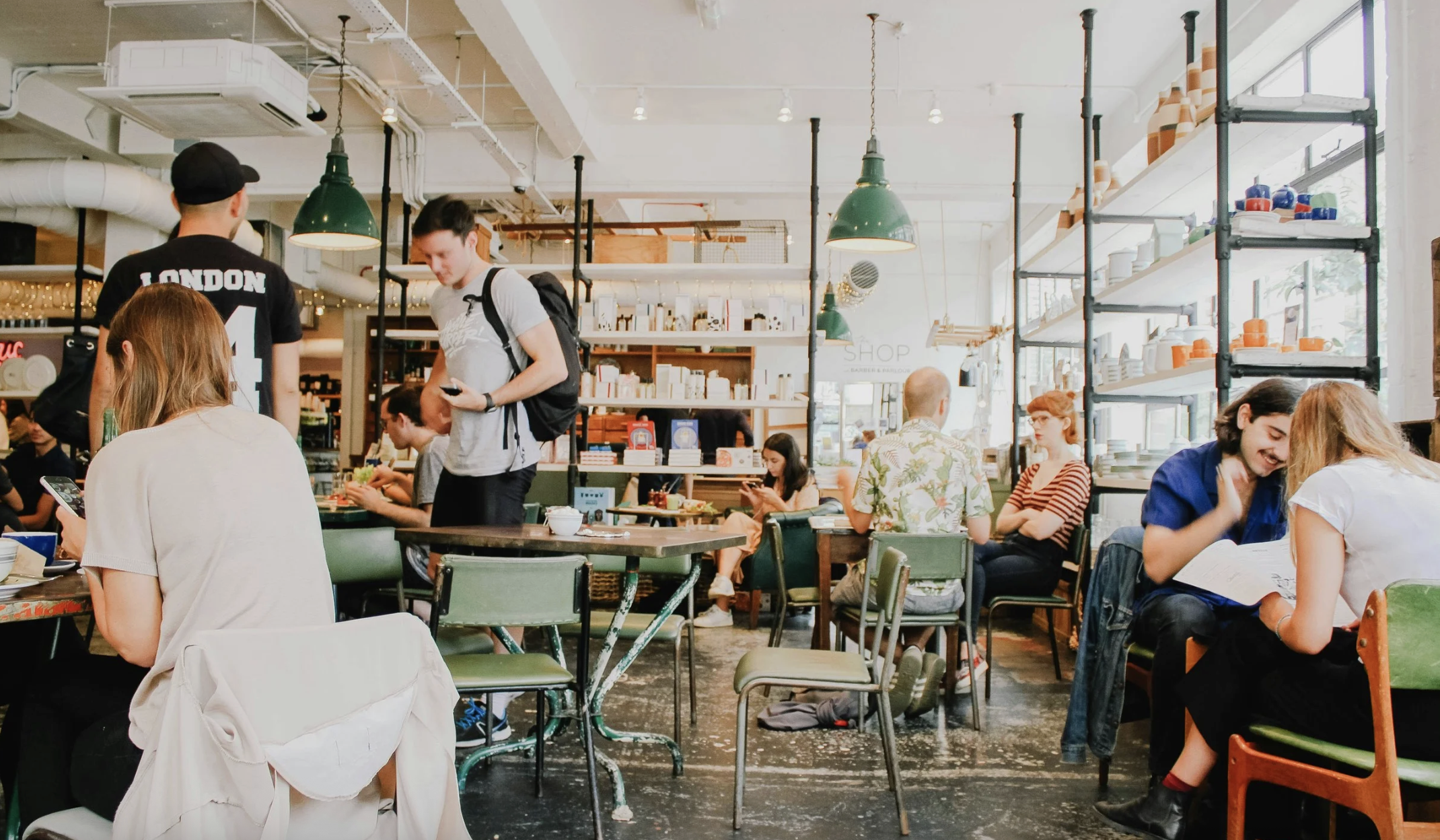 A busy café with people sitting at tables, some using their phones or reading. There are shelving units with dishes and decor, green pendant lights, and large windows letting in natural light.