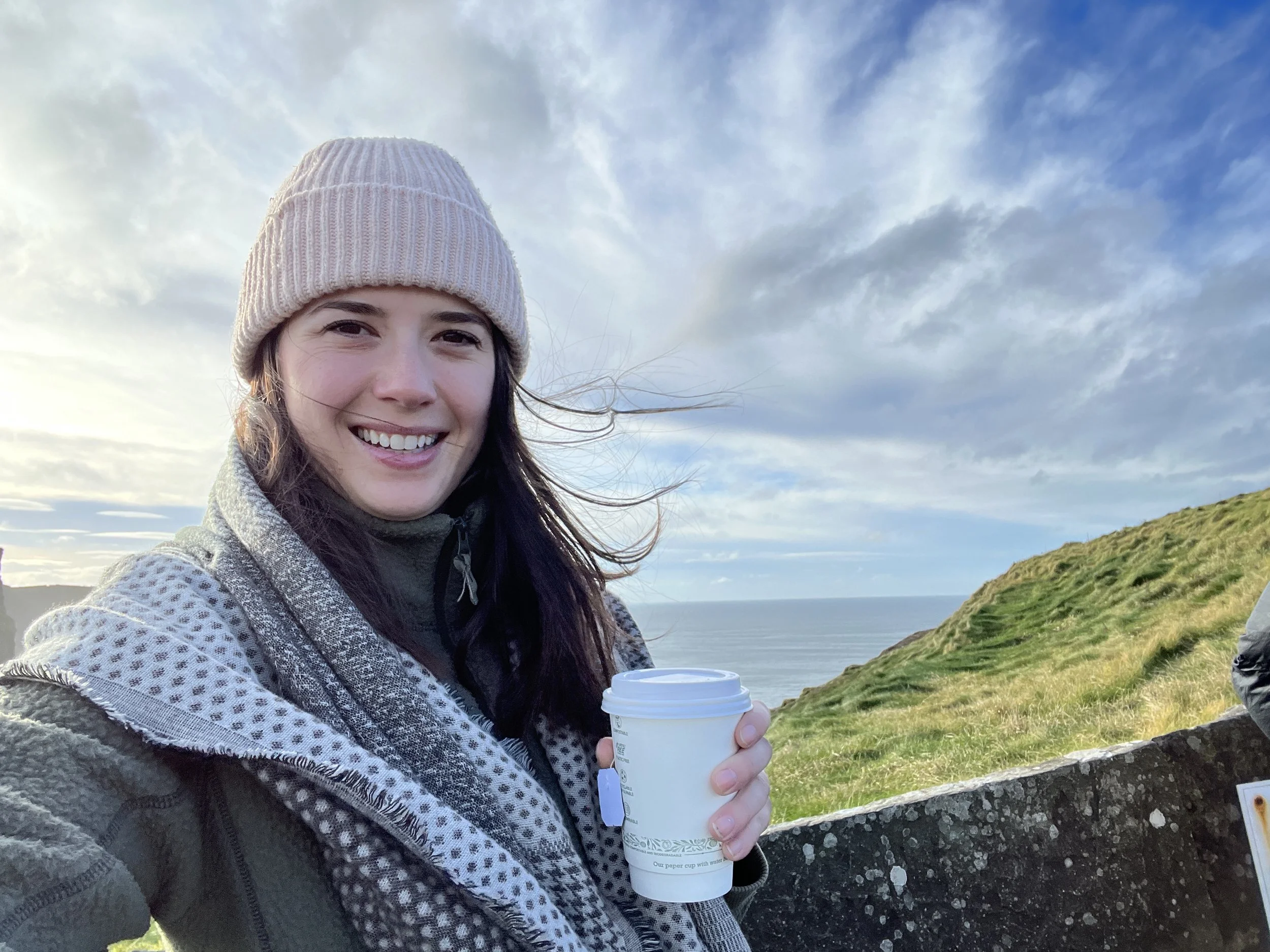 A smiling woman wearing a beige knit beanie and outdoor gear, holding a cup of coffee, standing near a grassy hill with the ocean and cloudy sky in the background.