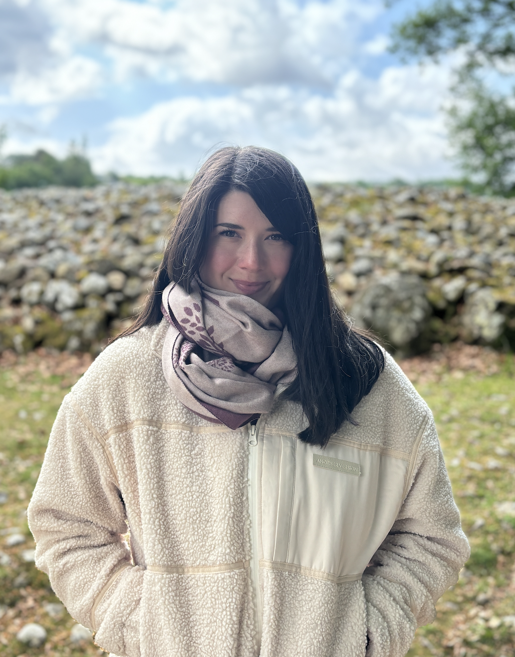 A woman with dark hair smiling outdoors, wearing a beige fuzzy jacket and a patterned scarf, standing in front of a rocky landscape with a partly cloudy sky.
