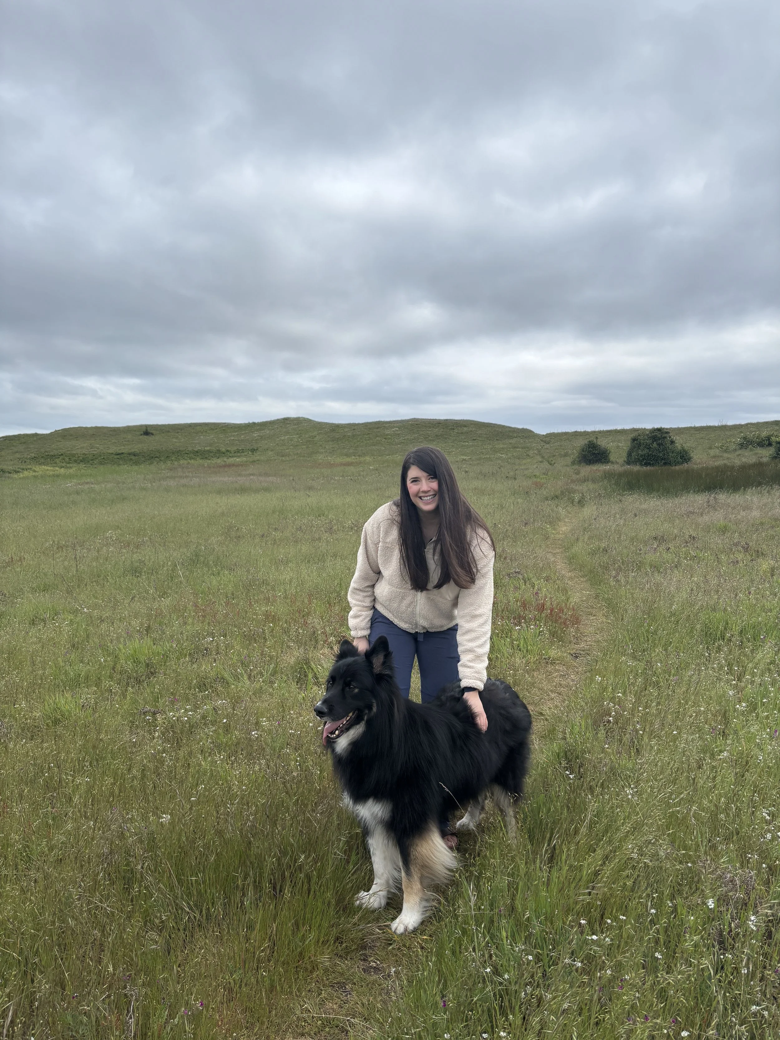 A woman with long dark hair smiling and standing in a grassy field with a black and white dog, under a cloudy sky.