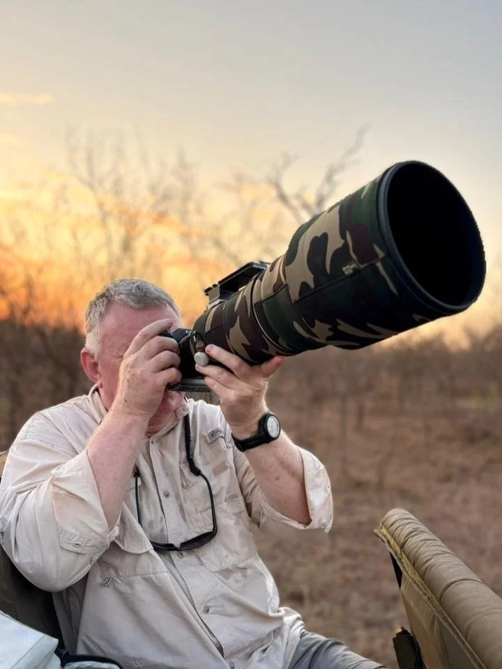 Man using a large camera with a camouflage lens outdoors during sunset or sunrise.
