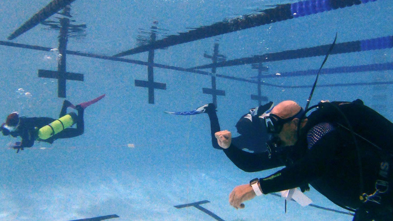 Two scuba divers underwater near a swimming pool wall with lane lines and cross markers.