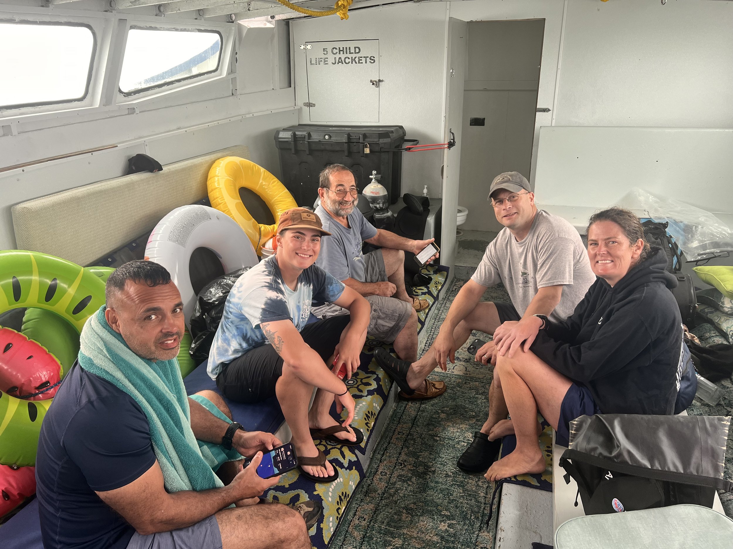 Six people sitting inside a boat cabin, with pool floats and beach gear, smiling and relaxing.