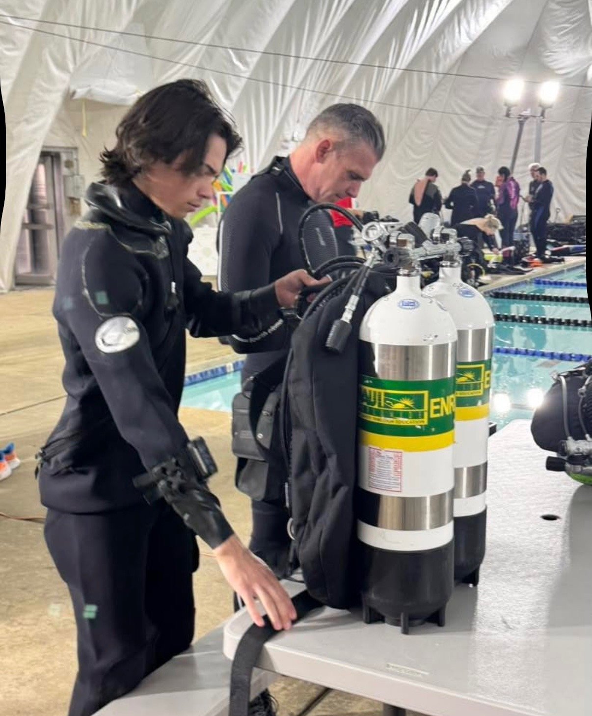 Two scuba divers standing near a swimming pool area, preparing their oxygen tanks, surrounded by other people.