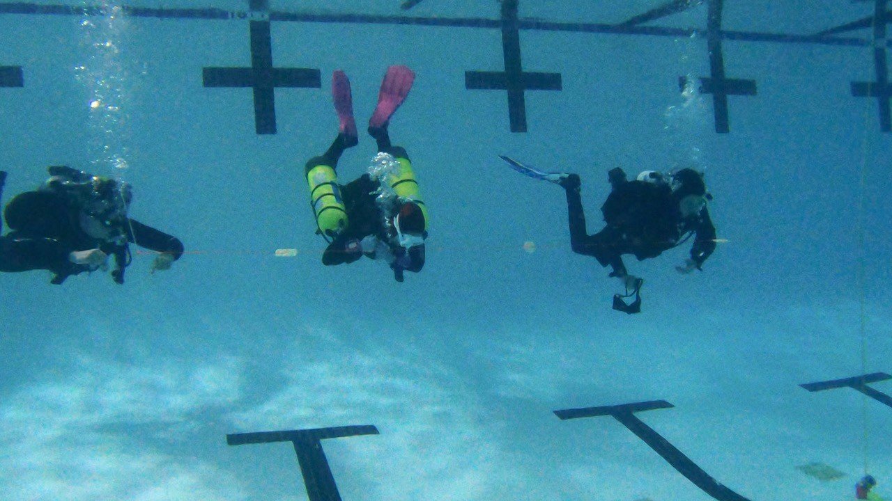 Three scuba divers underwater in a swimming pool, near lane markers.