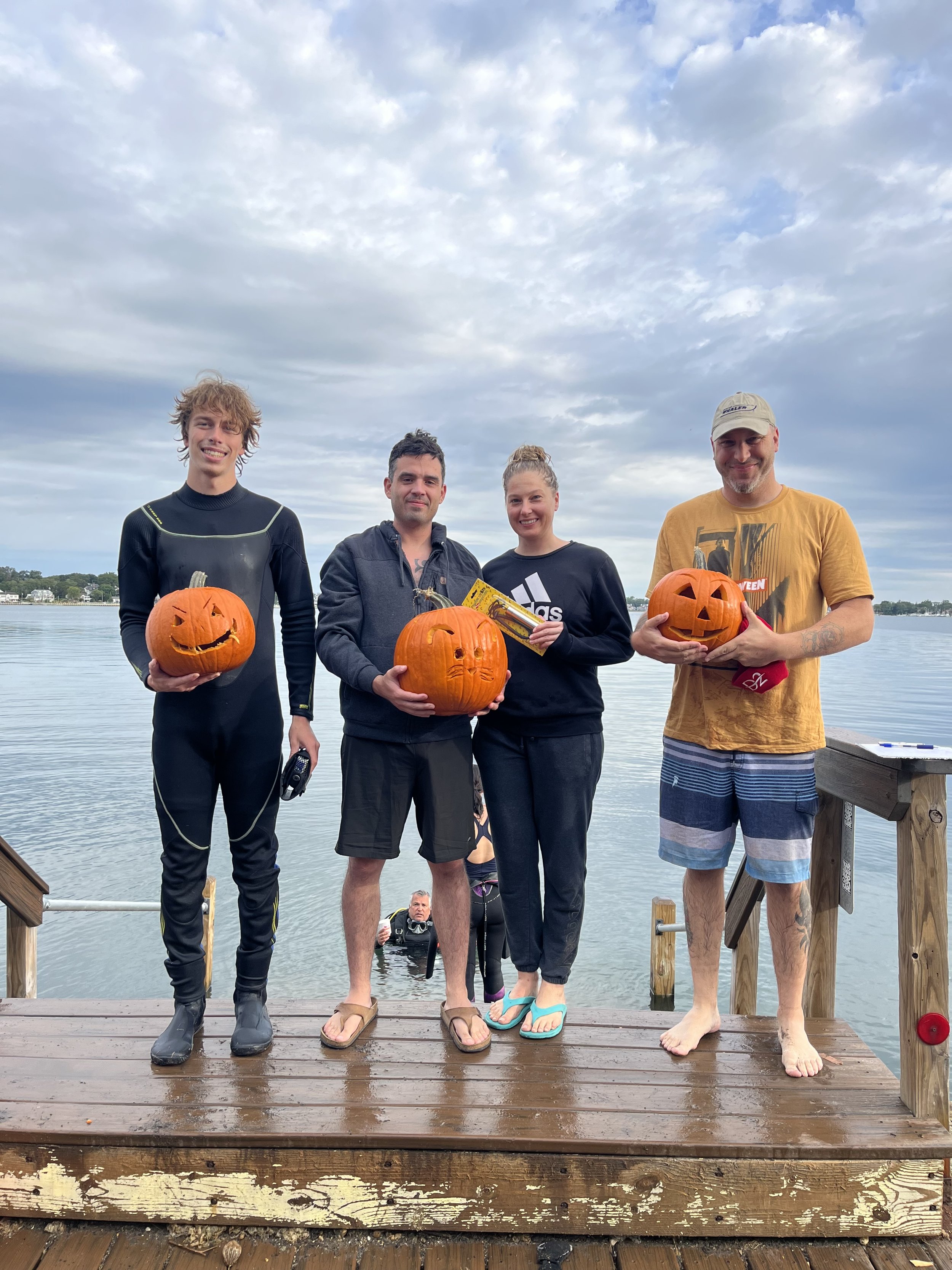 Four people standing on a dock by the water holding decorated pumpkins with carved faces. The sky is cloudy, and there is a person in the water behind them.