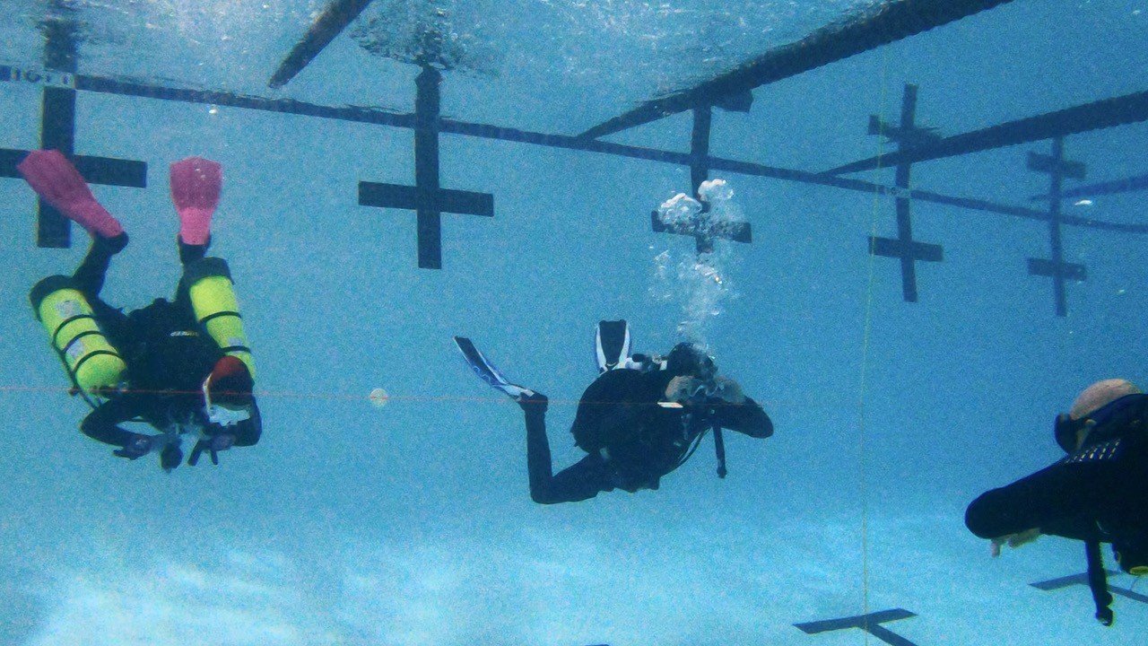 Two scuba divers underwater in a swimming pool, with one taking a photograph and the other looking at the camera, near lane markers.