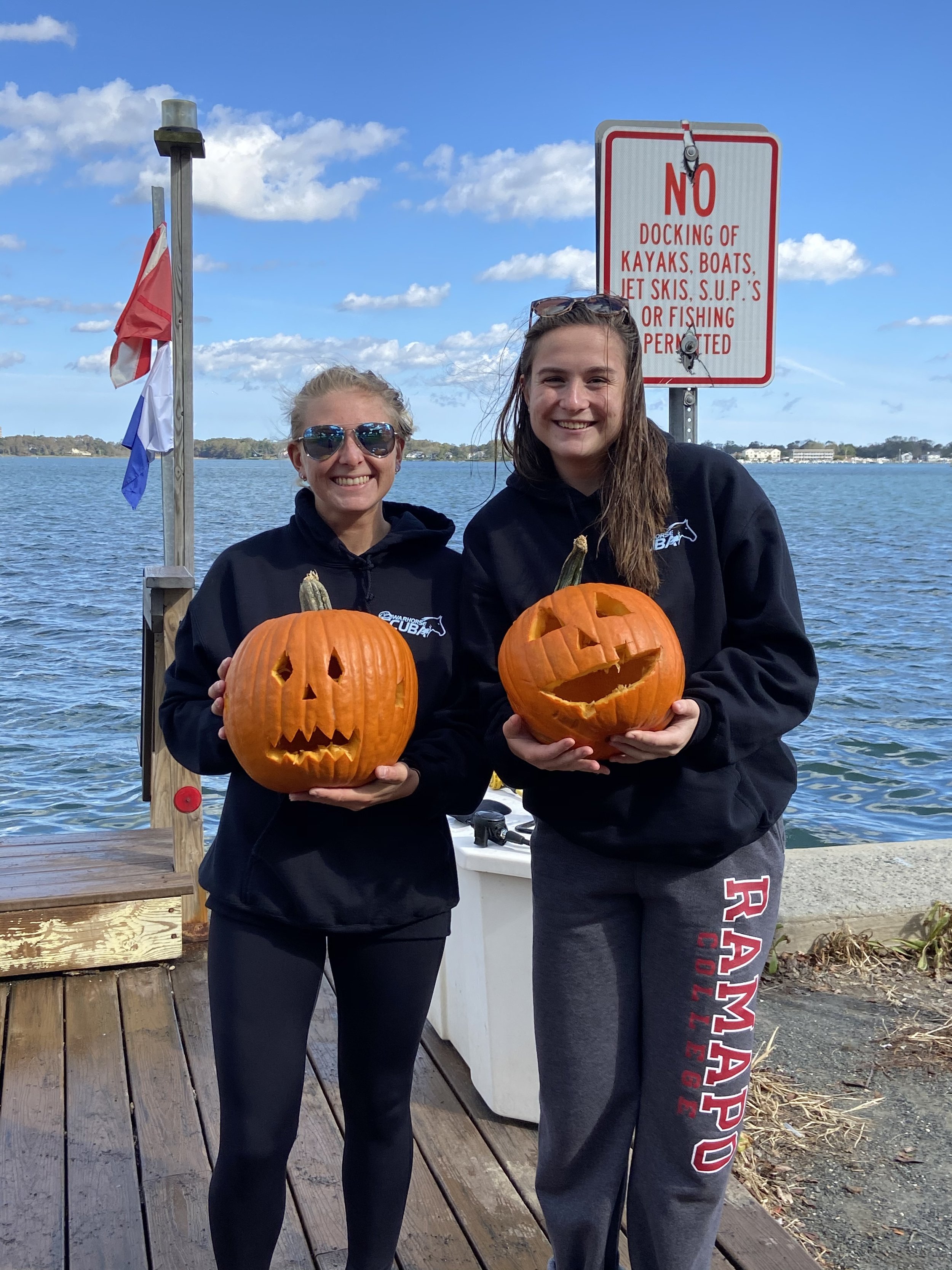 Two young women smiling and holding carved pumpkins by a lake, with a sign behind them indicating no docking or fishing allowed.