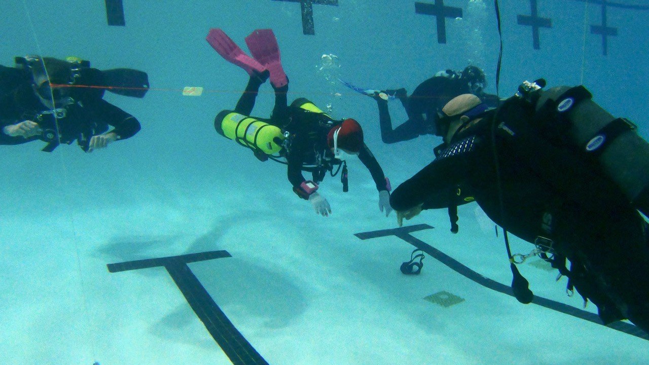 Group of divers underwater performing a training exercise with black pools and crosses on the pool floor.