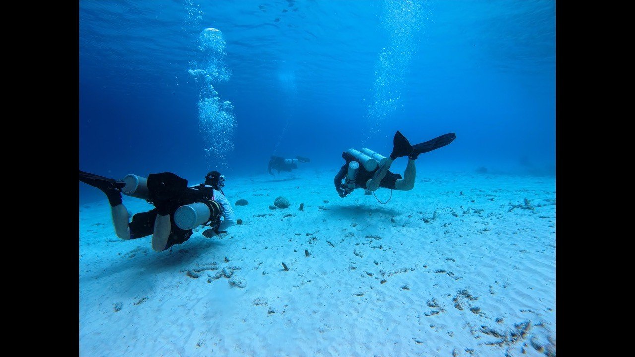 Two scuba divers swimming underwater in the ocean with sandy seafloor and blue water, surrounded by bubbles.