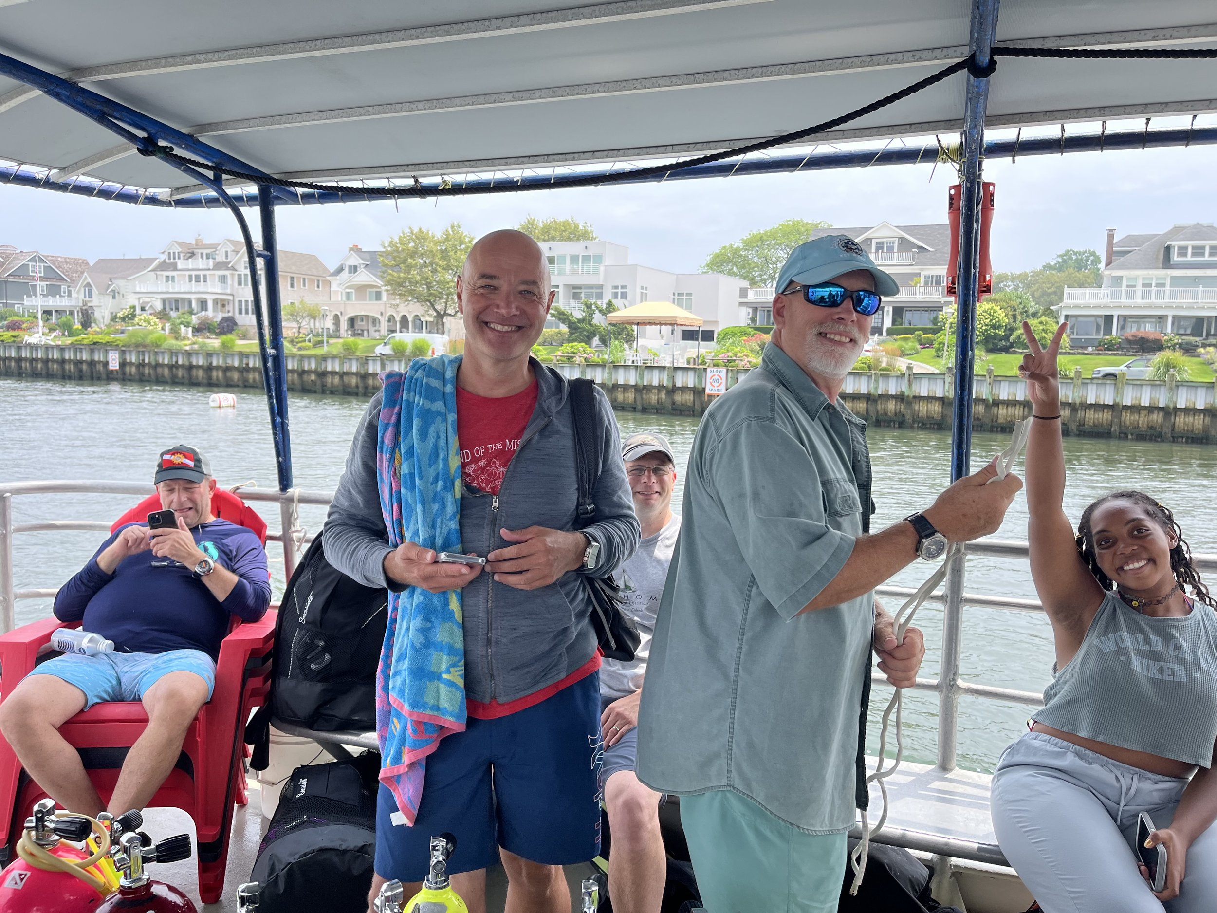 A group of people on a boat near a neighborhood waterfront, smiling and enjoying a sunny day with houses and trees in the background.