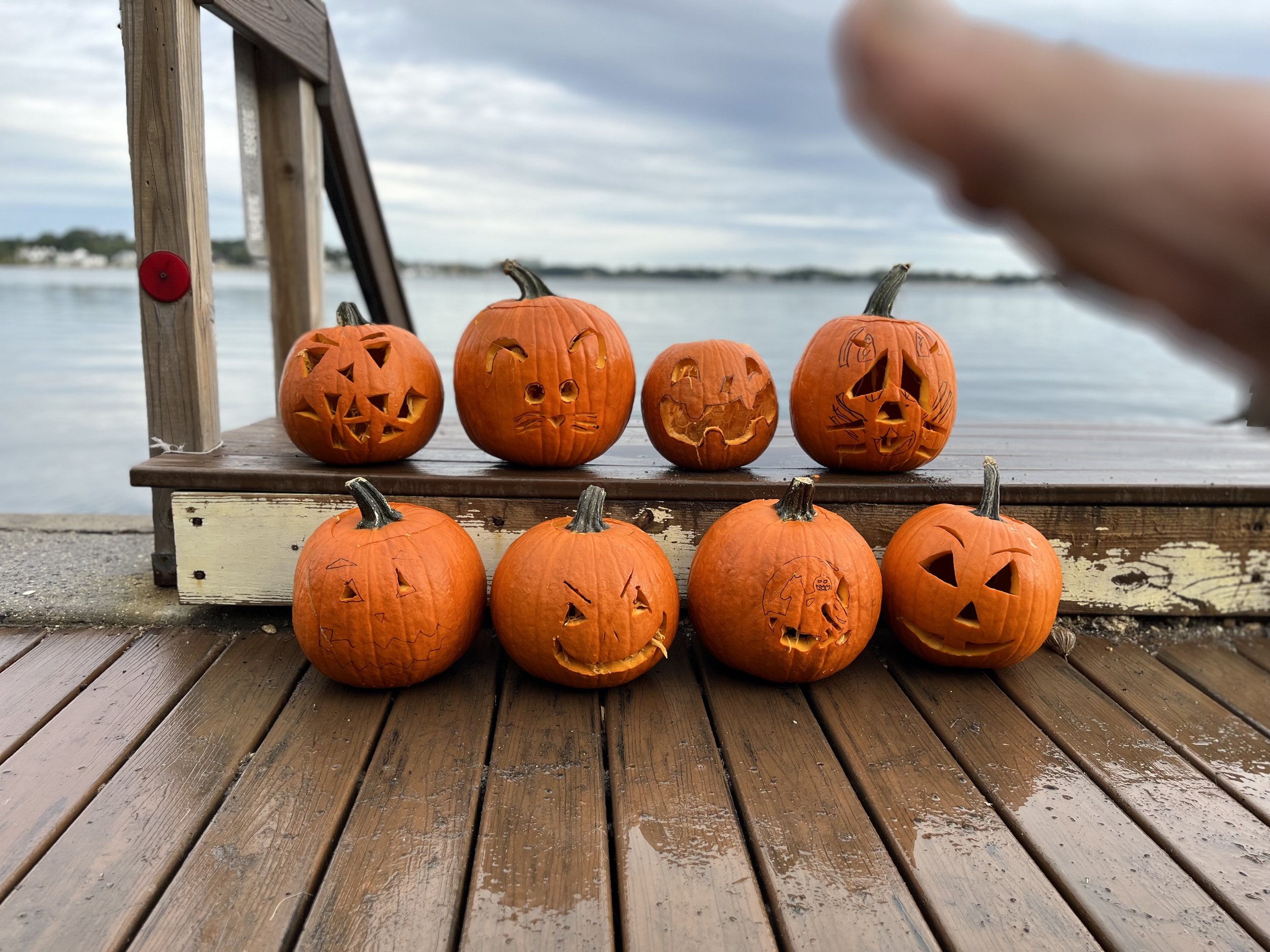 Seven carved pumpkins with various faces on a wooden dock near water, with a cloudy sky in the background.