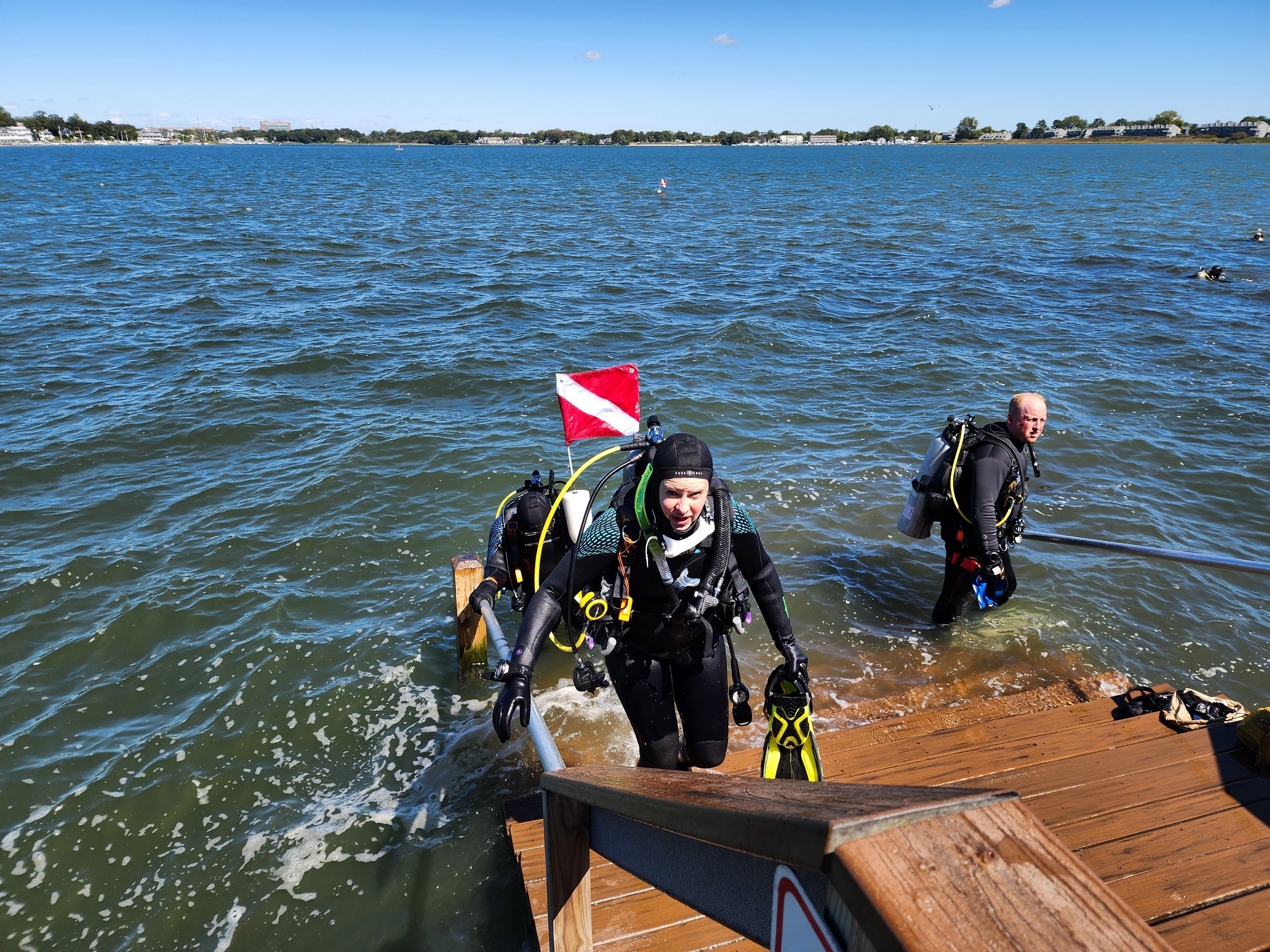 Two scuba divers emerging from the water at a wooden dock, wearing full gear including tanks, wetsuits, and masks, with a flag attached to one of the tanks, and a cityscape with trees and buildings in the background.
