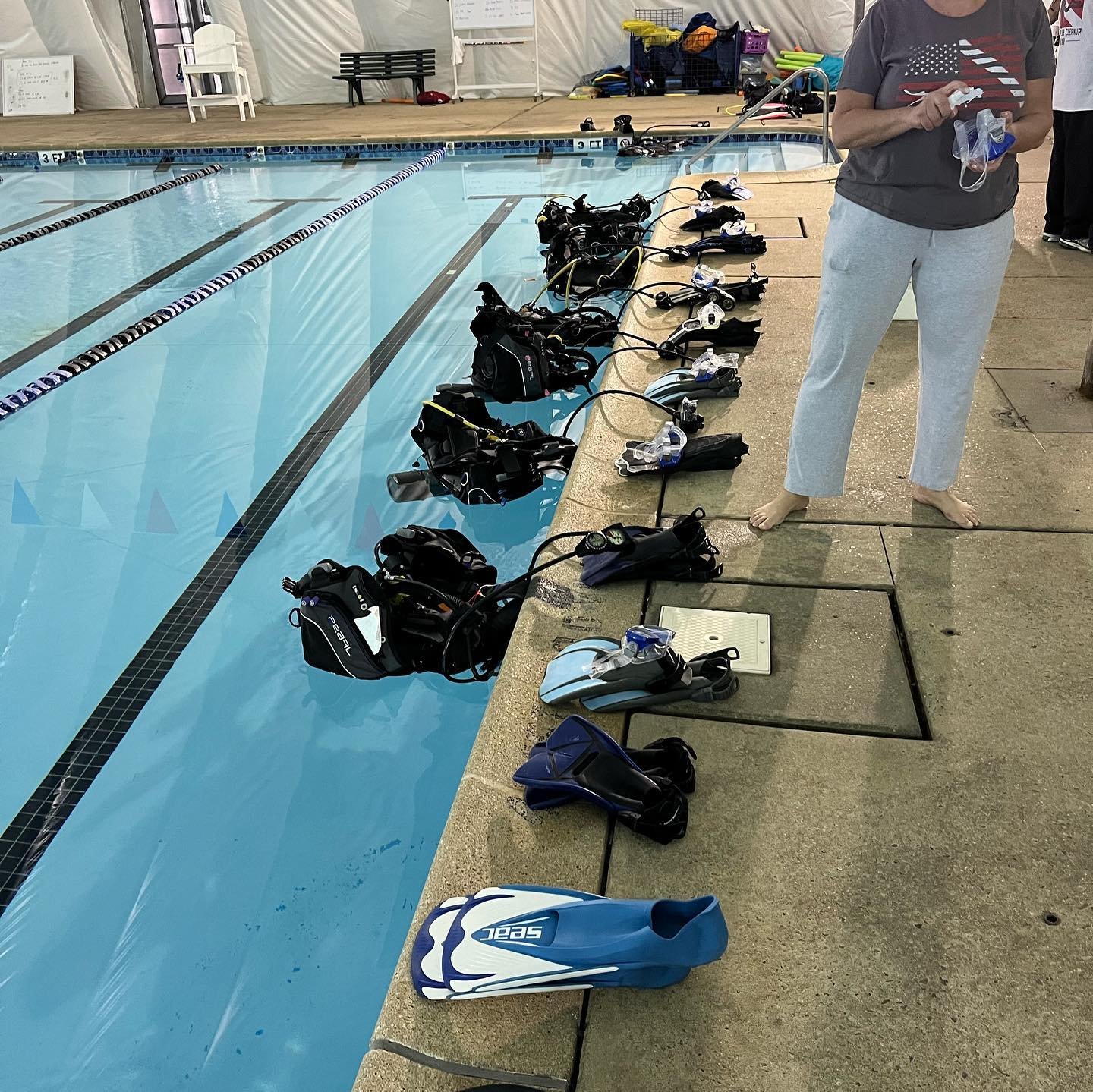 Diving equipment, including fins, masks, snorkels, and buoyancy compensators, arranged along the edge of an indoor swimming pool, with a person standing nearby holding more gear.