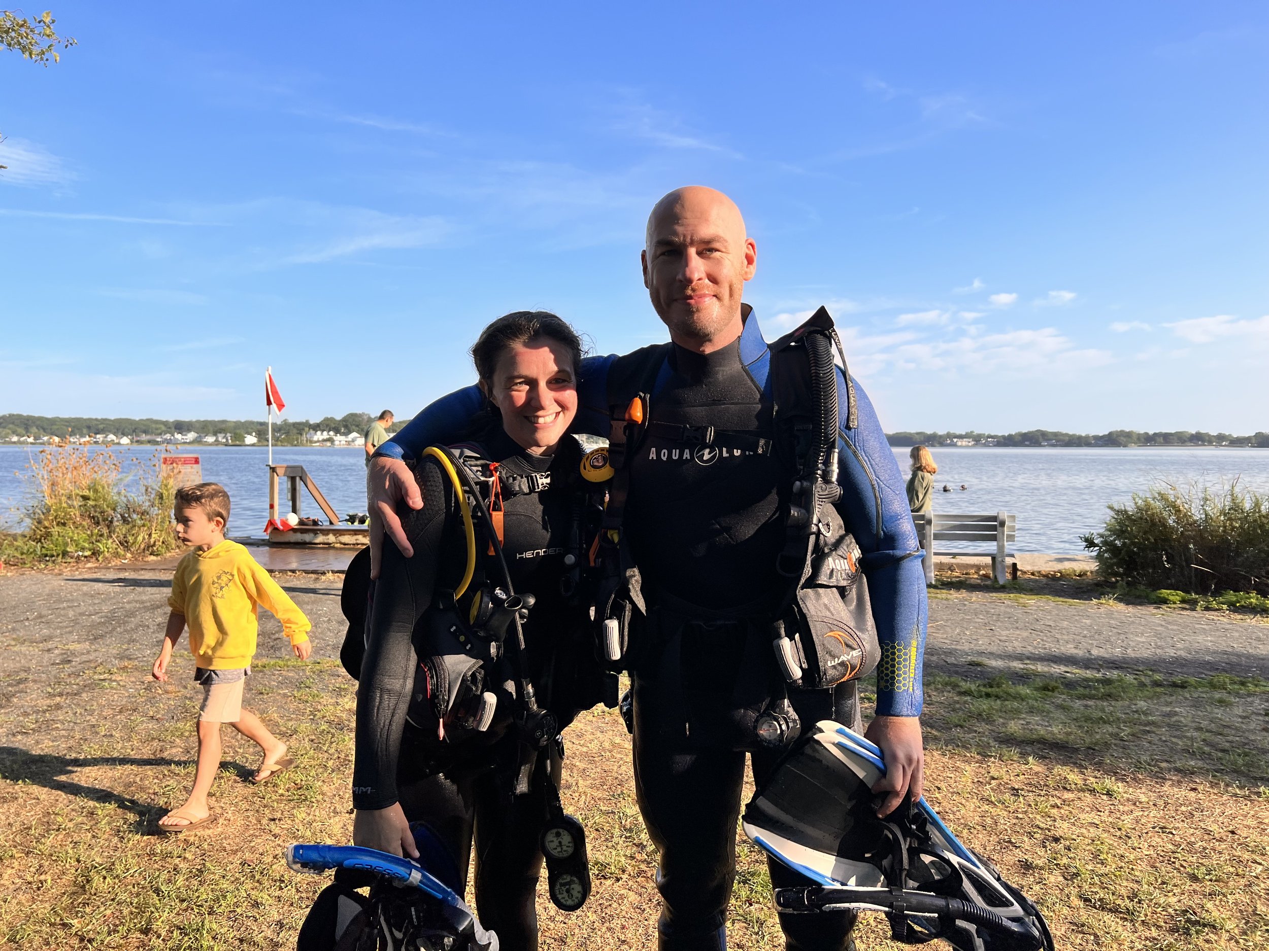 Two scuba divers with gear, smiling, standing near a lake during sunset, with children and a boat in the background.