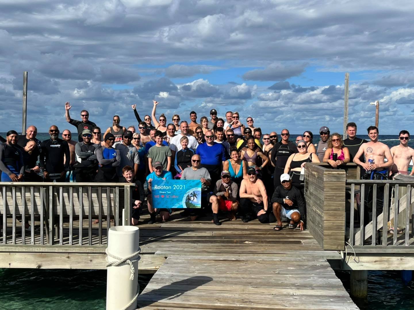 Group of people on a wooden dock by the ocean, holding a blue banner that reads "Roatan 2021 Diver's Trip," with partly cloudy sky overhead.