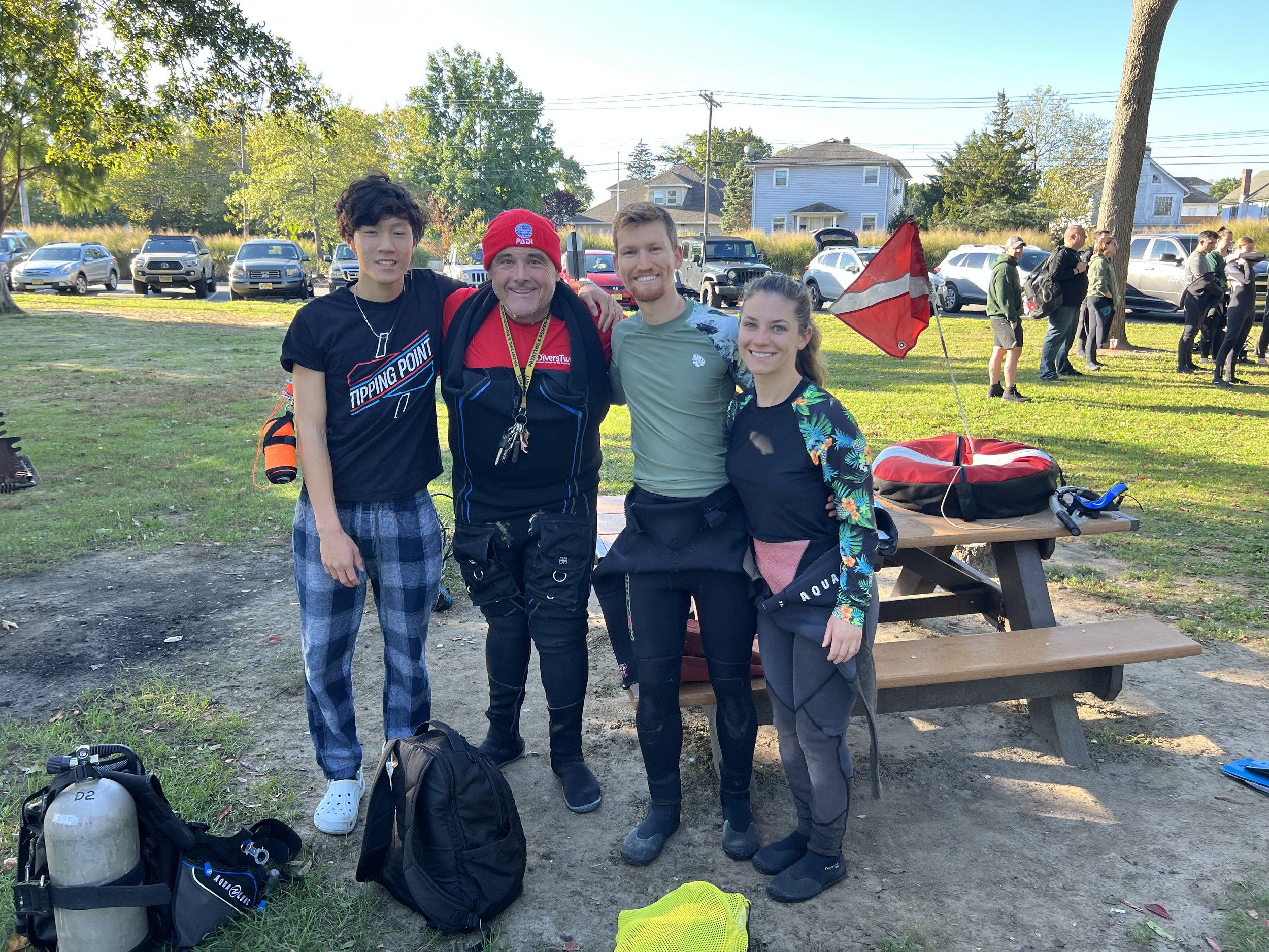 Four people standing outdoors in a park, smiling, with diving gear and a red flag nearby, with parked cars and trees in the background.