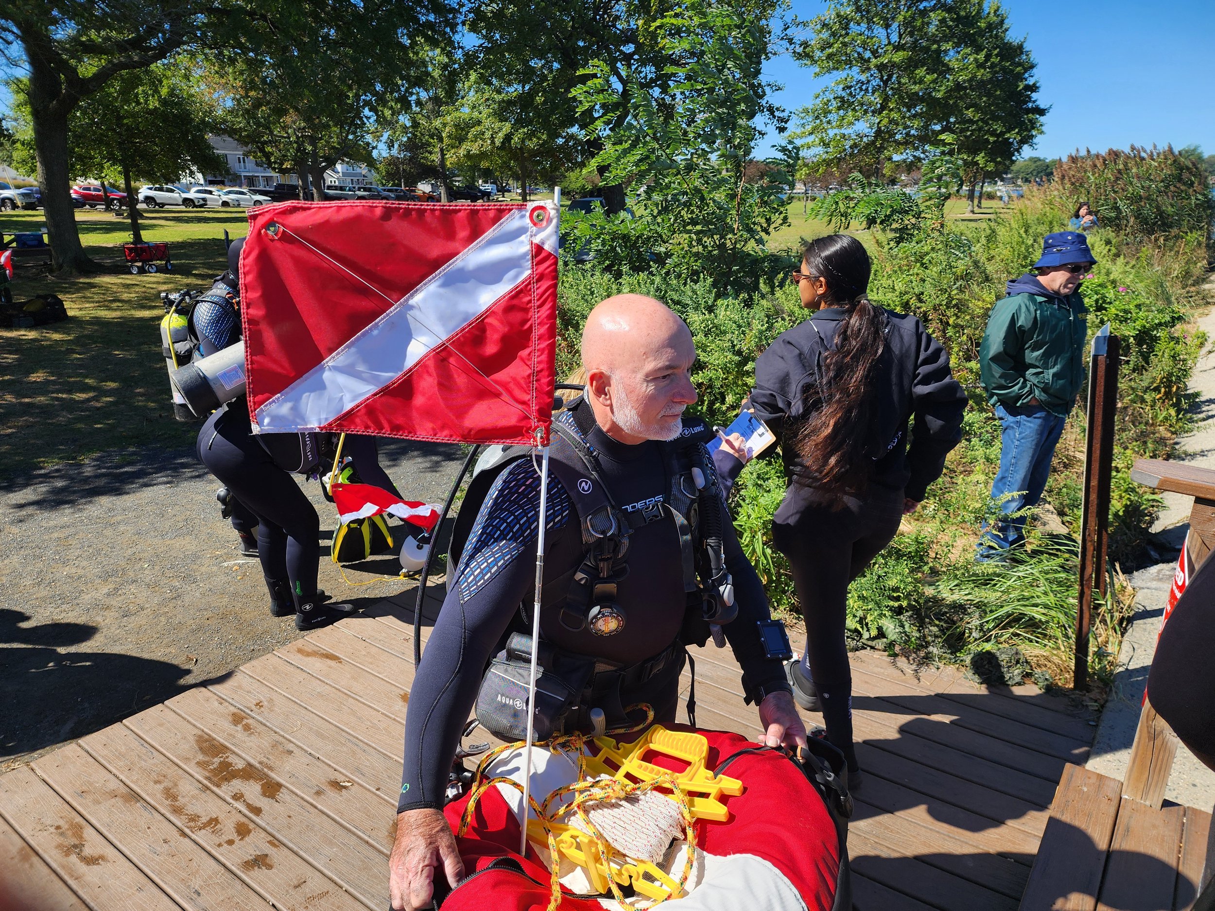 A man with a white beard and bald head sitting on a wooden dock, dressed in a wetsuit, with training or safety gear on his lap, outdoors on a sunny day. Multiple people are present in the background among trees and greenery.
