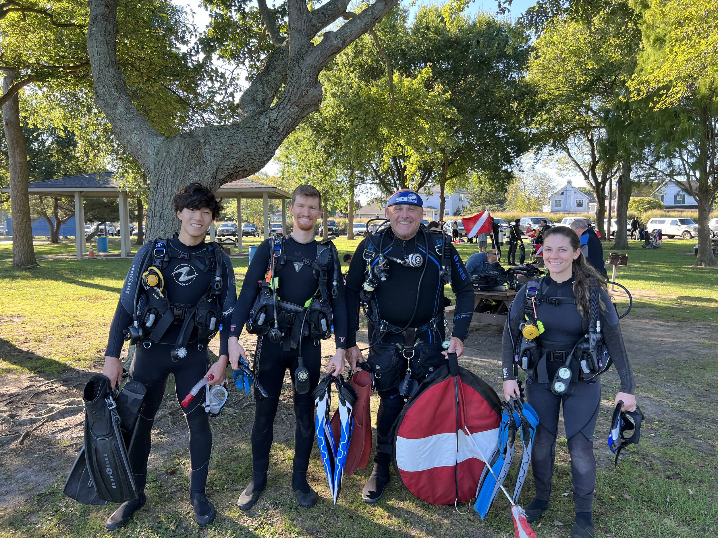 Four scuba divers in black wetsuits standing outdoors under a large tree, holding diving gear, with parks and other people in the background.