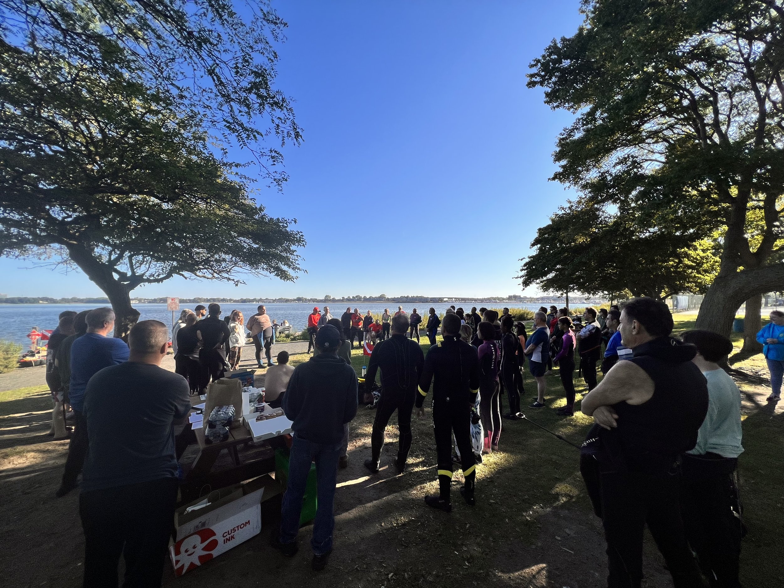 Group of people gathered by a waterfront, near trees under a clear blue sky.