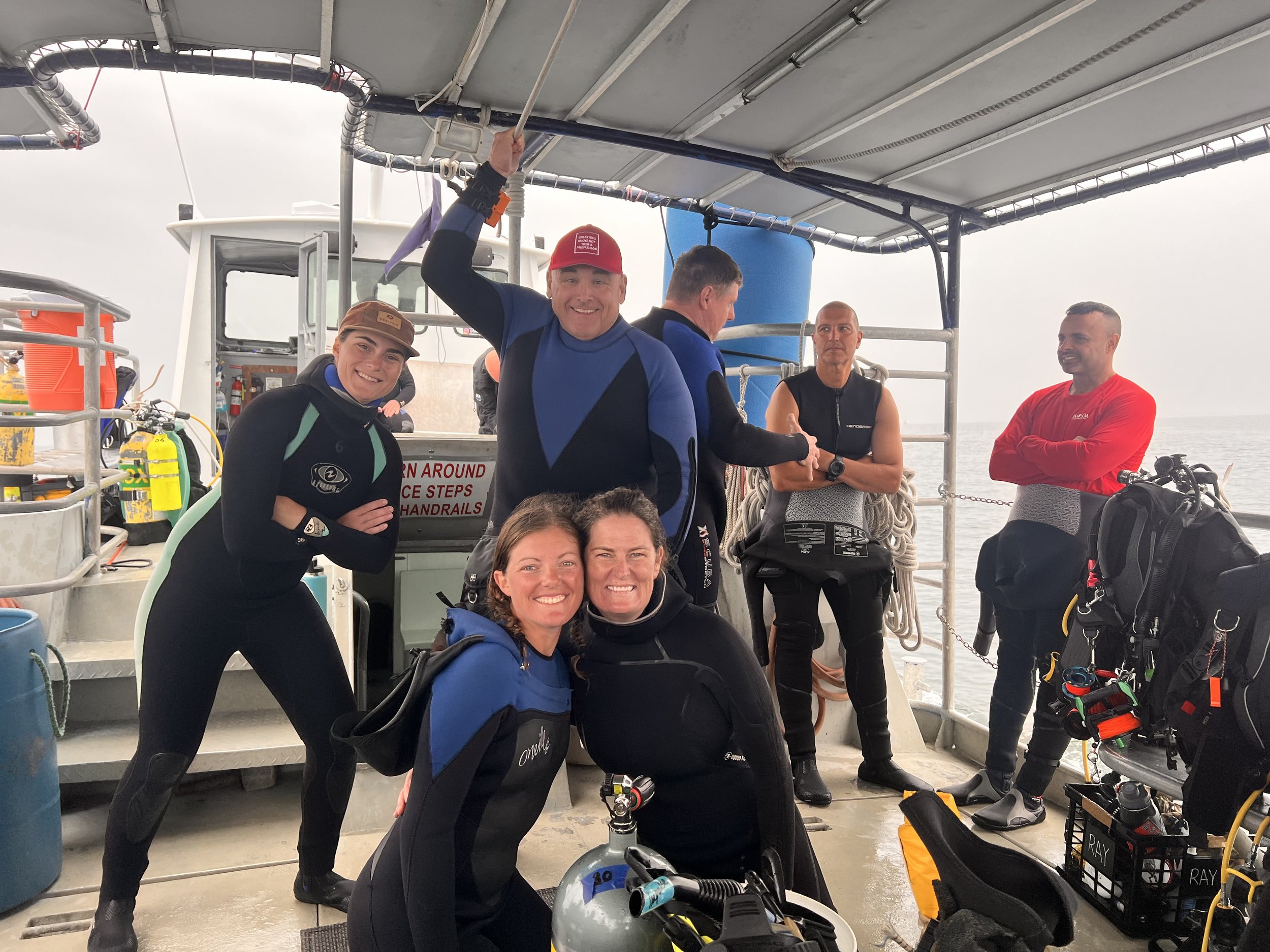 Group of people in wetsuits on a boat preparing for a scuba diving adventure, some smiling at the camera, with diving gear and equipment around them.