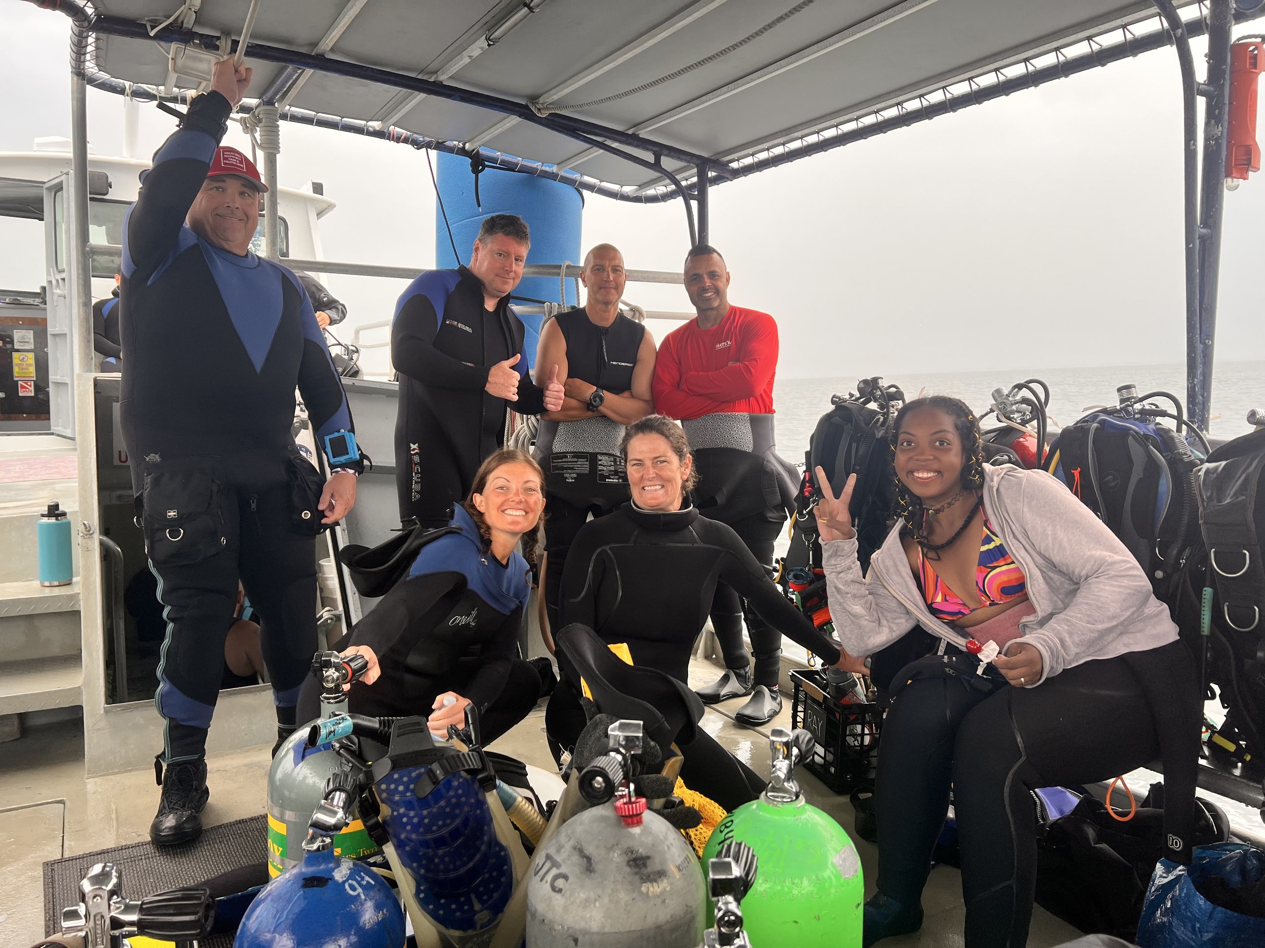 Group of divers posing on boat with scuba gear and underwater tanks, smiling and making peace signs.