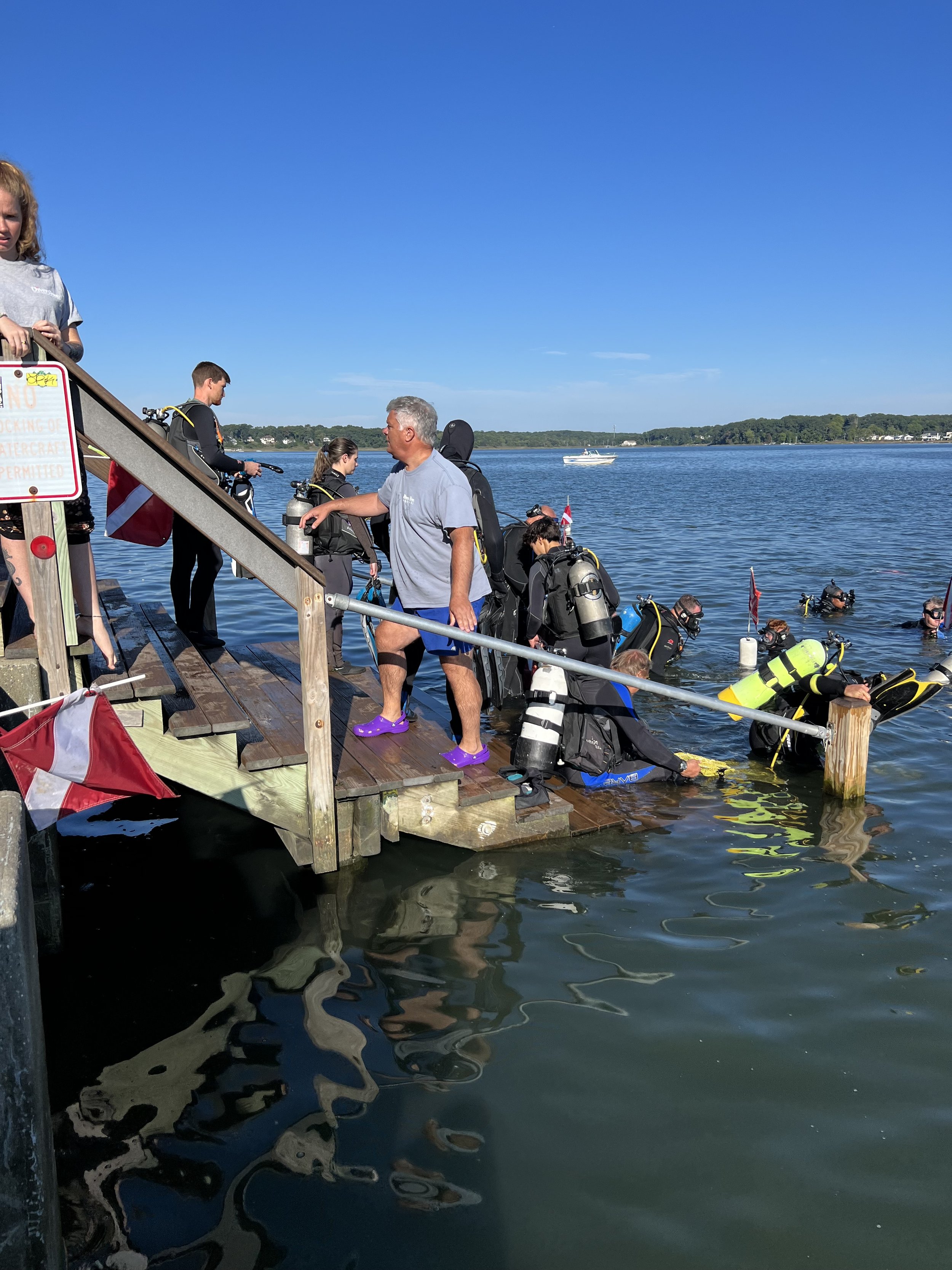 Diver training session with people in wetsuits and scuba gear on a wooden dock by the water, some are entering the water, and a boat is visible in the background.