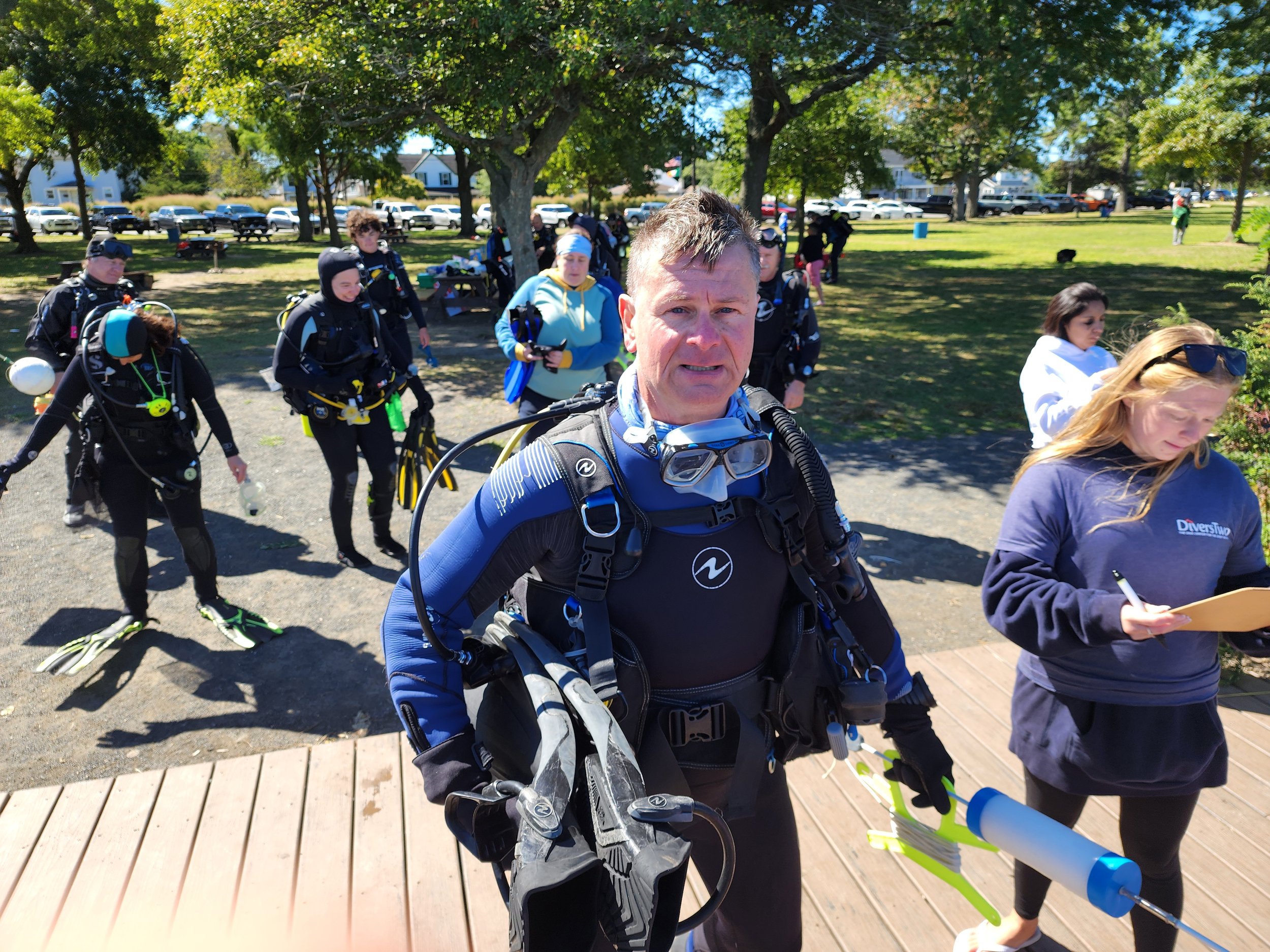 Group of scuba divers preparing for a dive at a park, with a man in the foreground holding a mask and scuba gear, and others in the background wearing wetsuits and holding diving equipment.