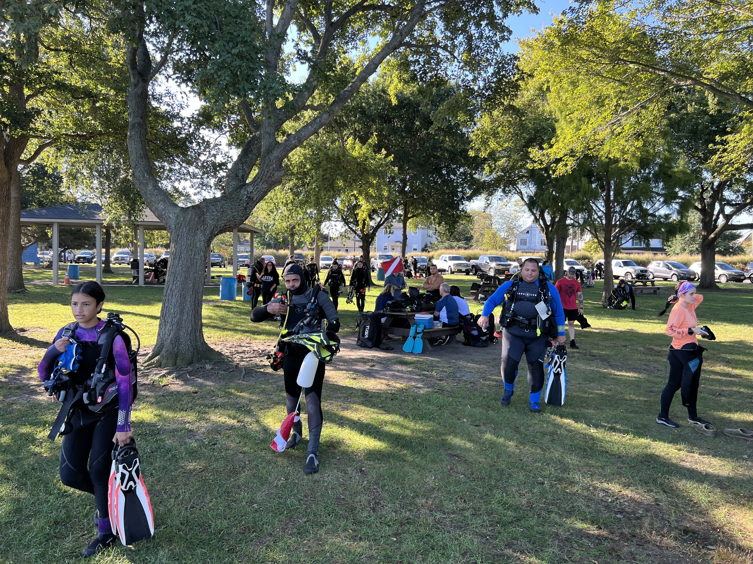 People preparing for a kayaking trip in a park with trees, picnic tables, and parked cars in the background.
