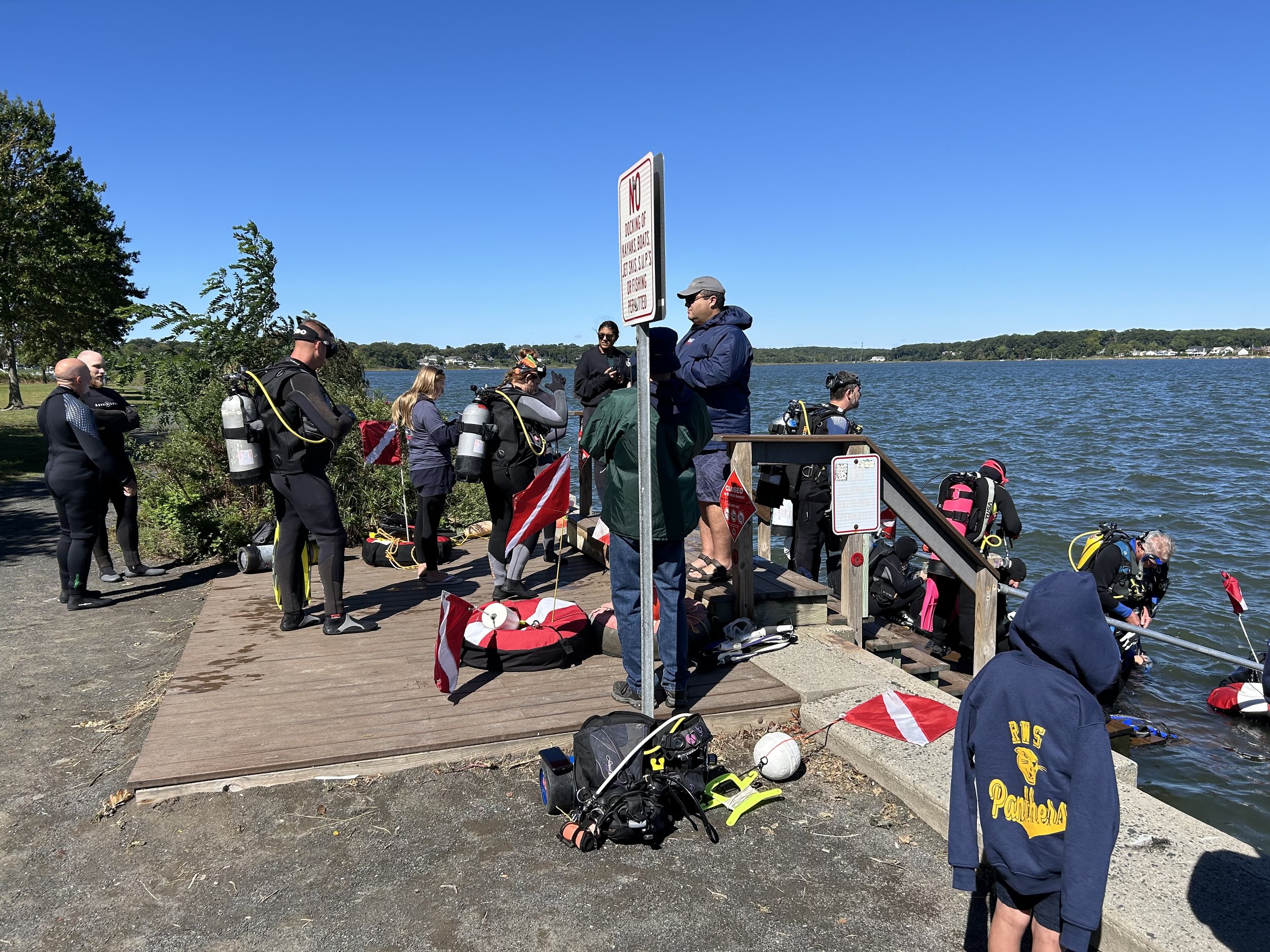 Group of people preparing for scuba diving on a wooden dock by a lake under a clear blue sky