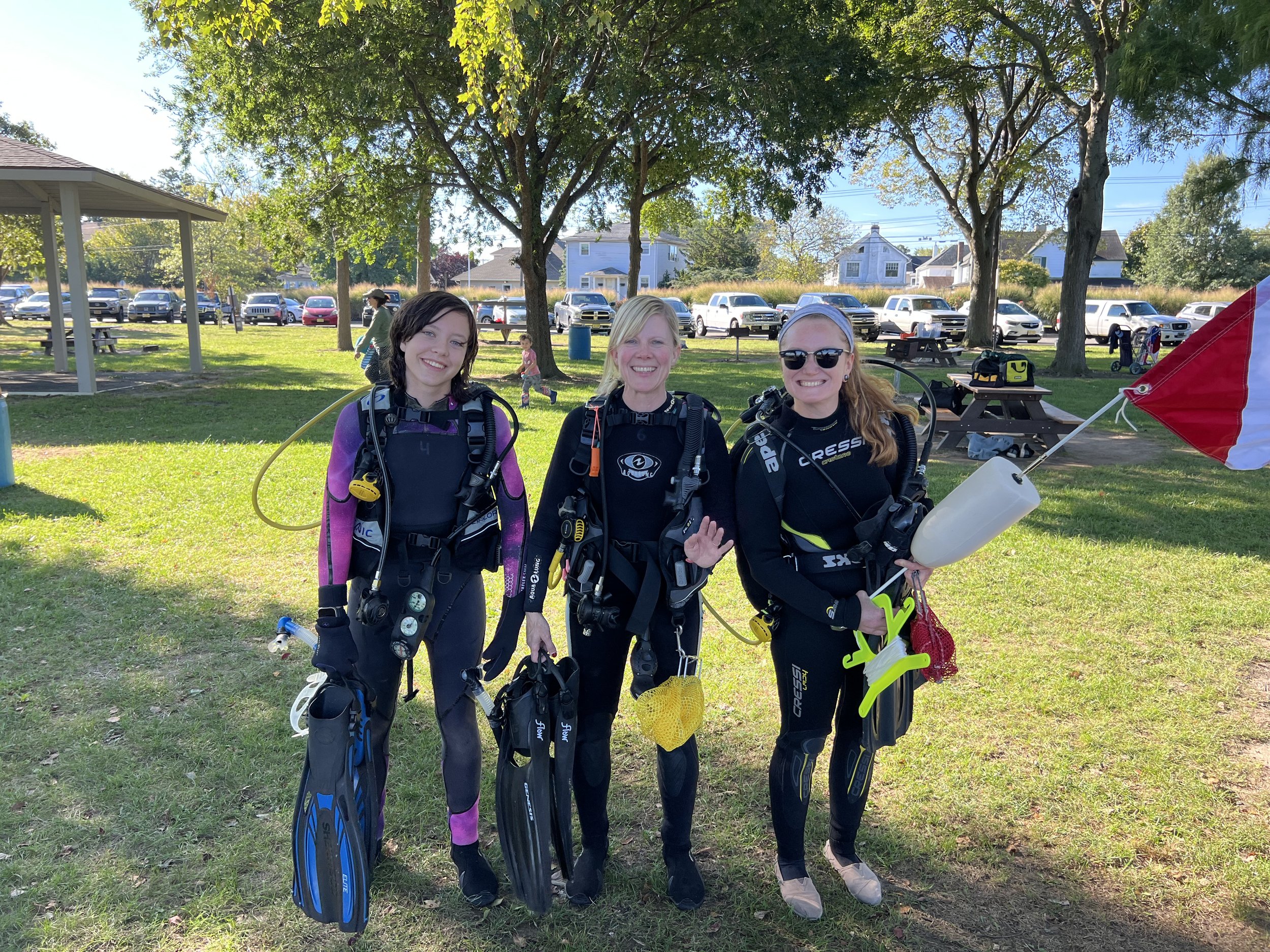 Three women in scuba diving gear standing on grass, smiling at the camera, with trees, picnic tables, and parked cars in the background.