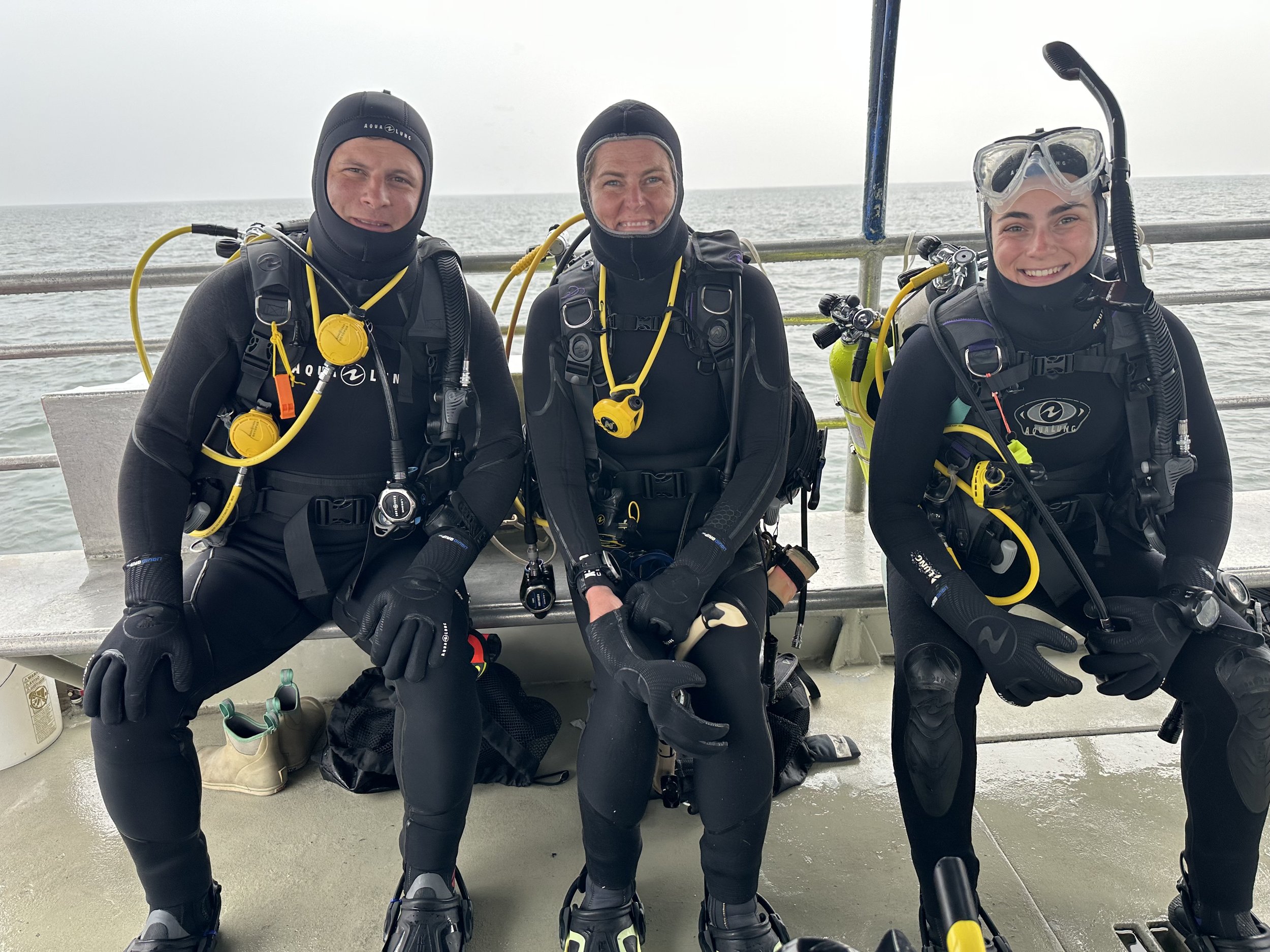 Three scuba divers in wetsuits and gear sitting on a boat with the ocean in the background.