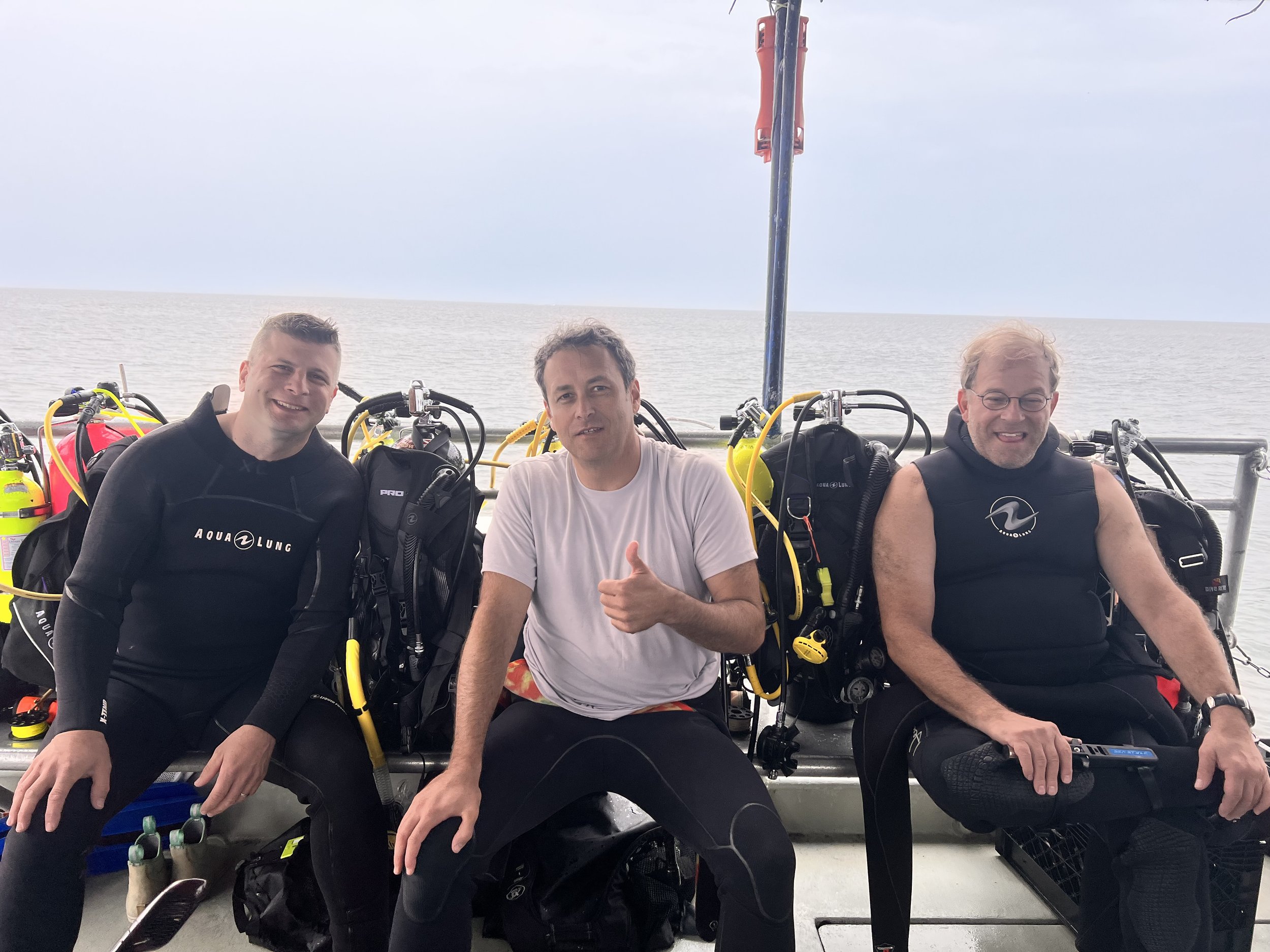 Three men sitting on a boat with scuba diving gear, smiling, with an ocean in the background.