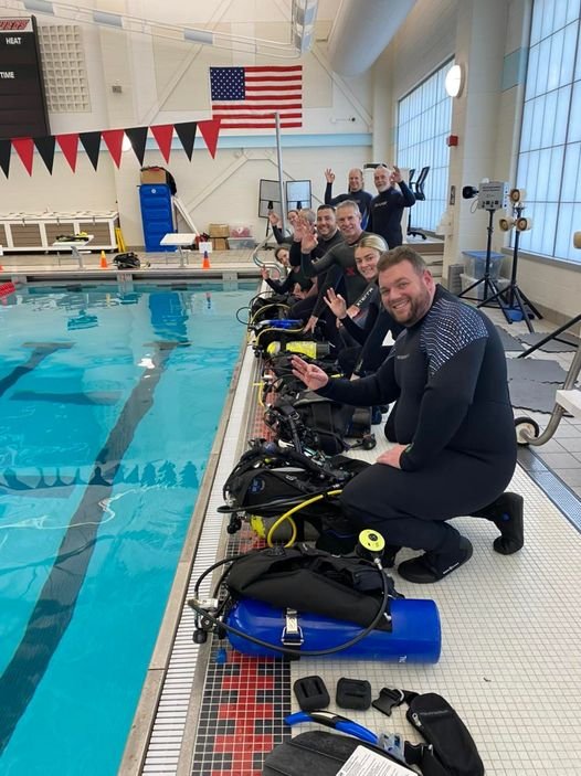 Group of people in wetsuits sitting on underwater scooters at an indoor swimming pool, smiling and waving at the camera with an American flag hanging on the wall in the background.