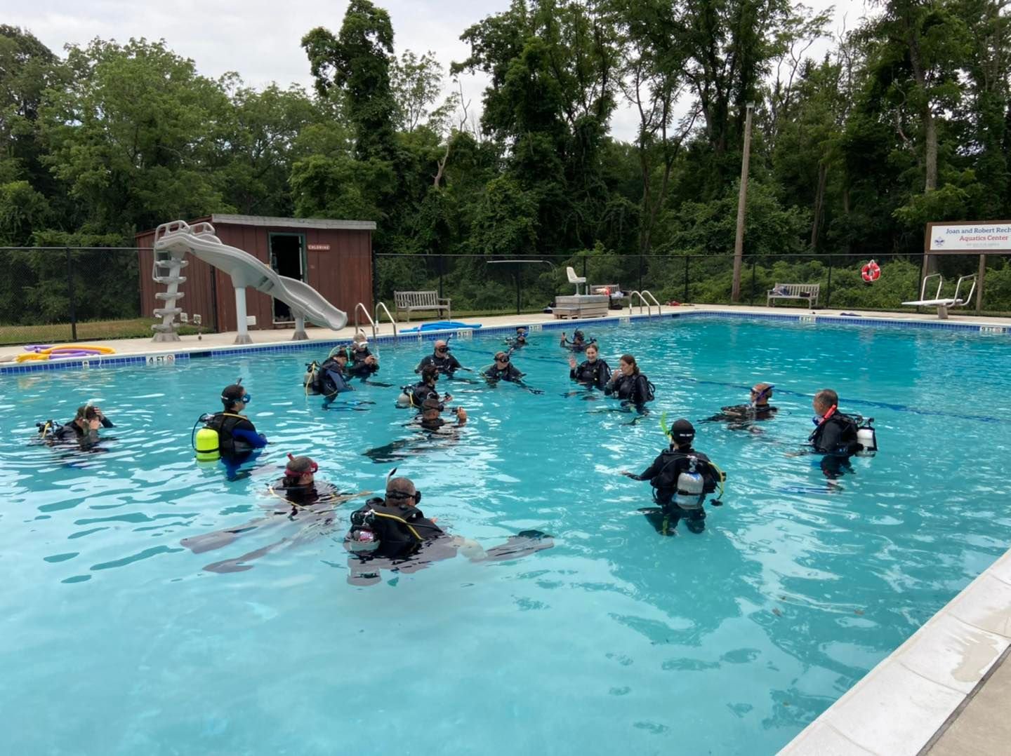 Group of people in scuba diving gear participating in a scuba training session in an outdoor swimming pool surrounded by trees.
