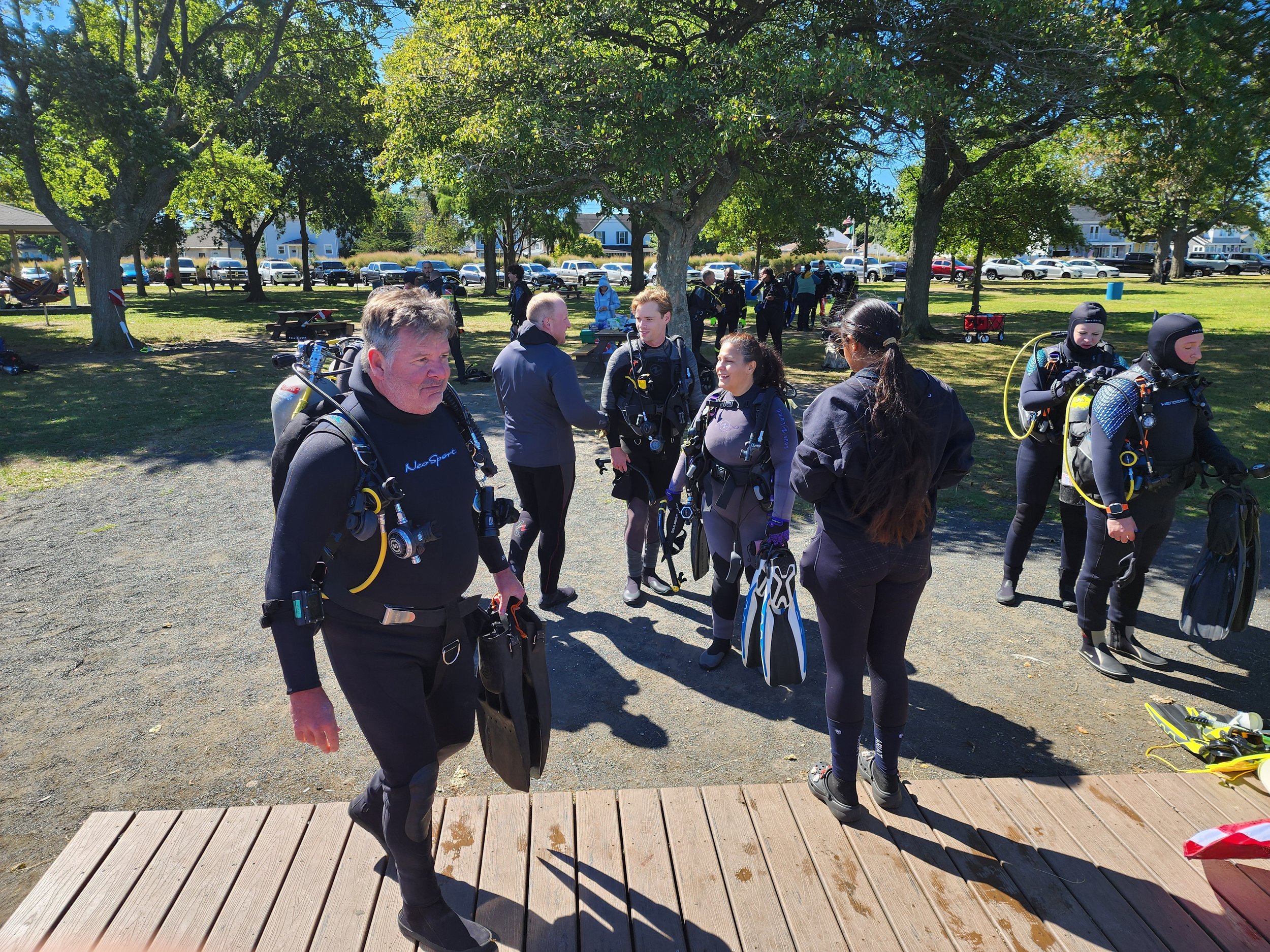 Group of people in scuba diving gear preparing for a dive in a park with trees and parked cars in the background.