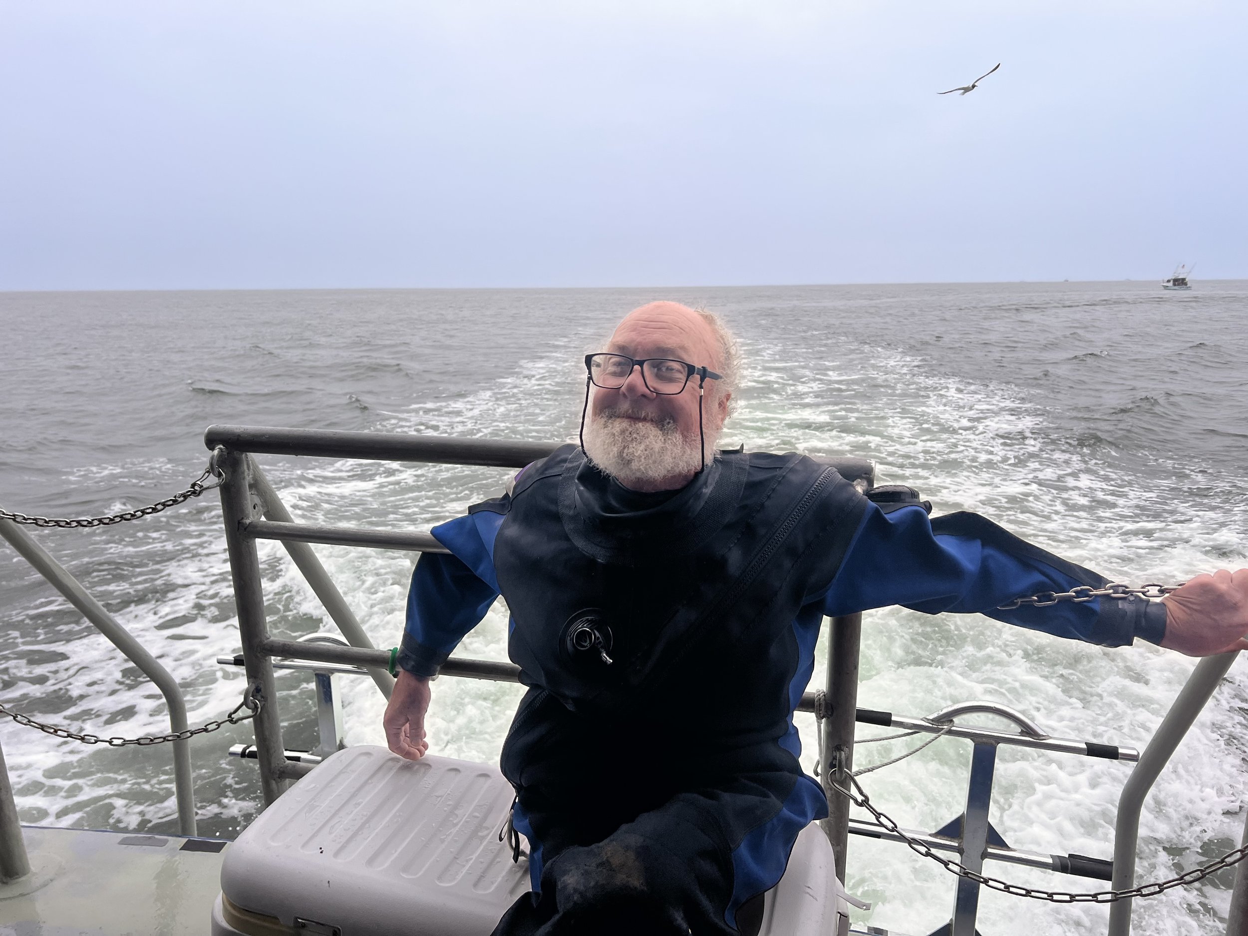 An elderly man with glasses and a beard smiling on a boat, holding onto the railing, with the open ocean and a seagull flying overhead in the background.