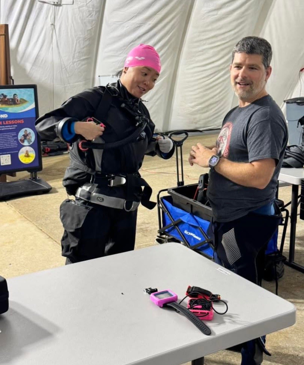 Two people inside a tent, one wearing scuba gear and the other in casual clothing, talking to each other.