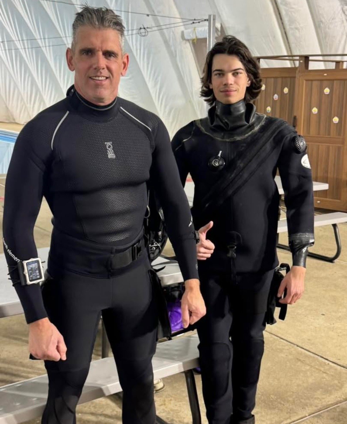 Two men in black athletic or dive suits standing indoors, one older and one younger, with a bench and white tent walls in the background.