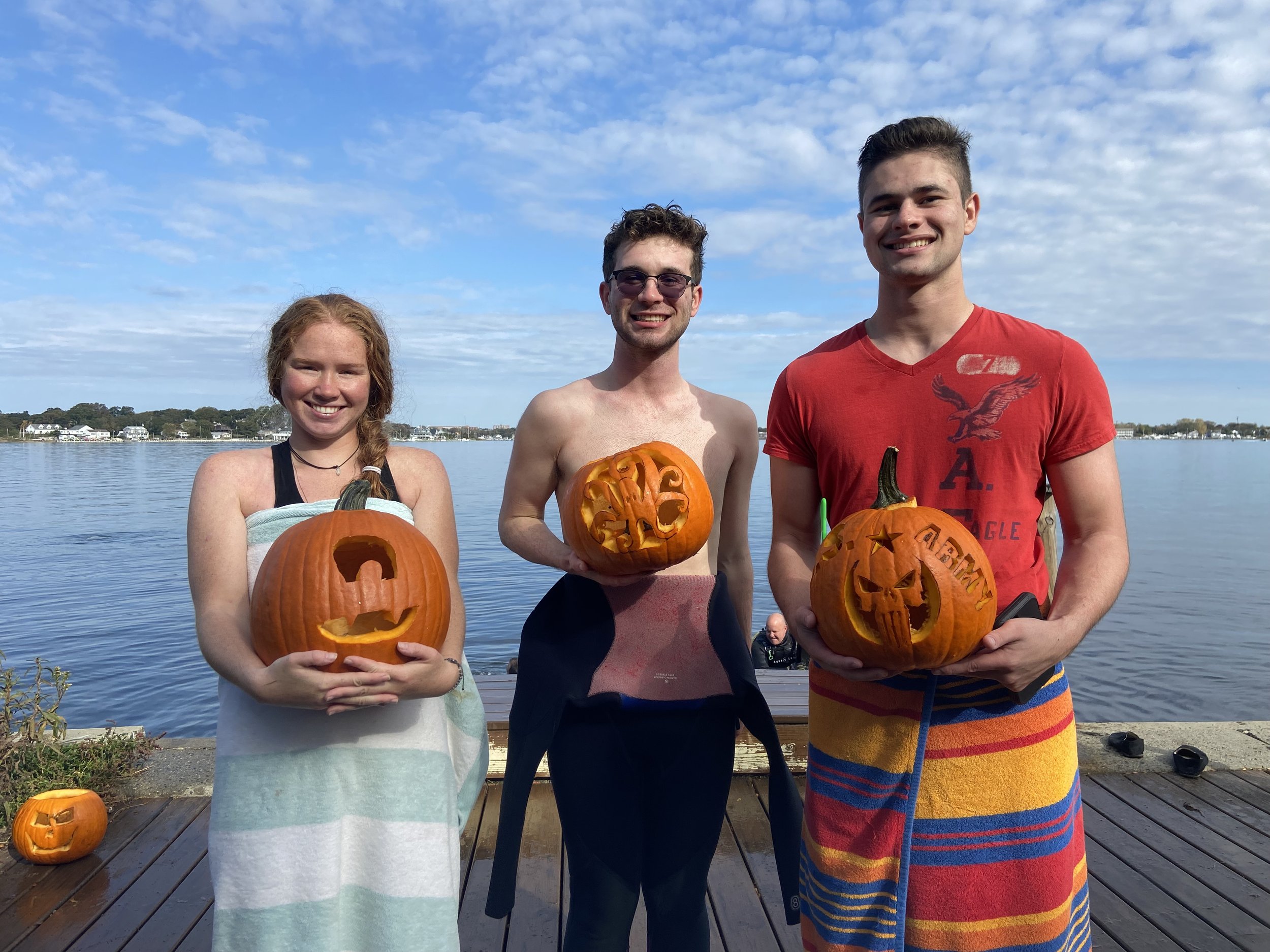 Three young adults holding carved Halloween pumpkins on a dock by a lake with a partly cloudy sky.