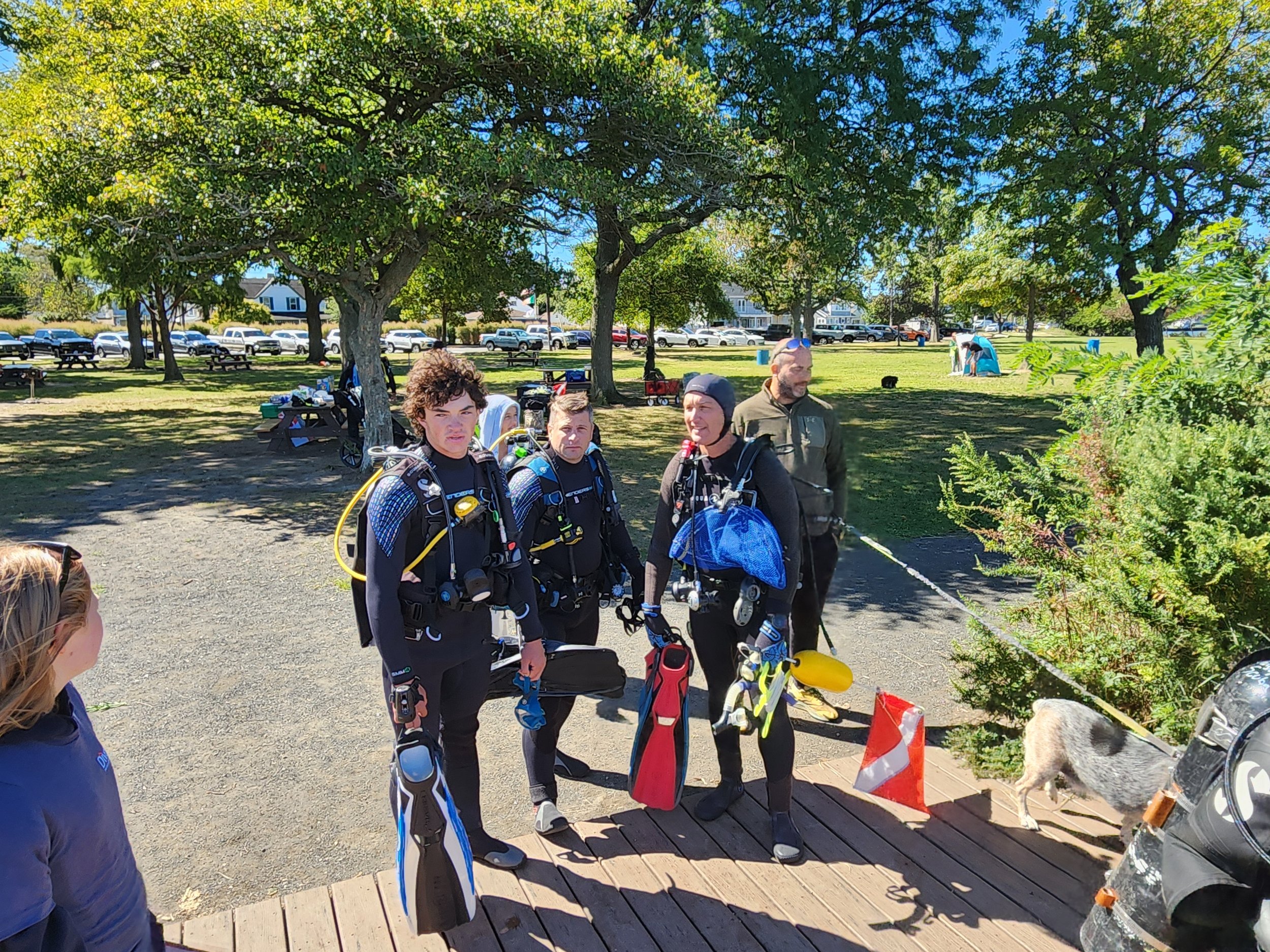 Group of four people in scuba diving gear standing on a wooden dock in a park, with trees, grass, and parked cars in the background, and a dog near the right side of the image.