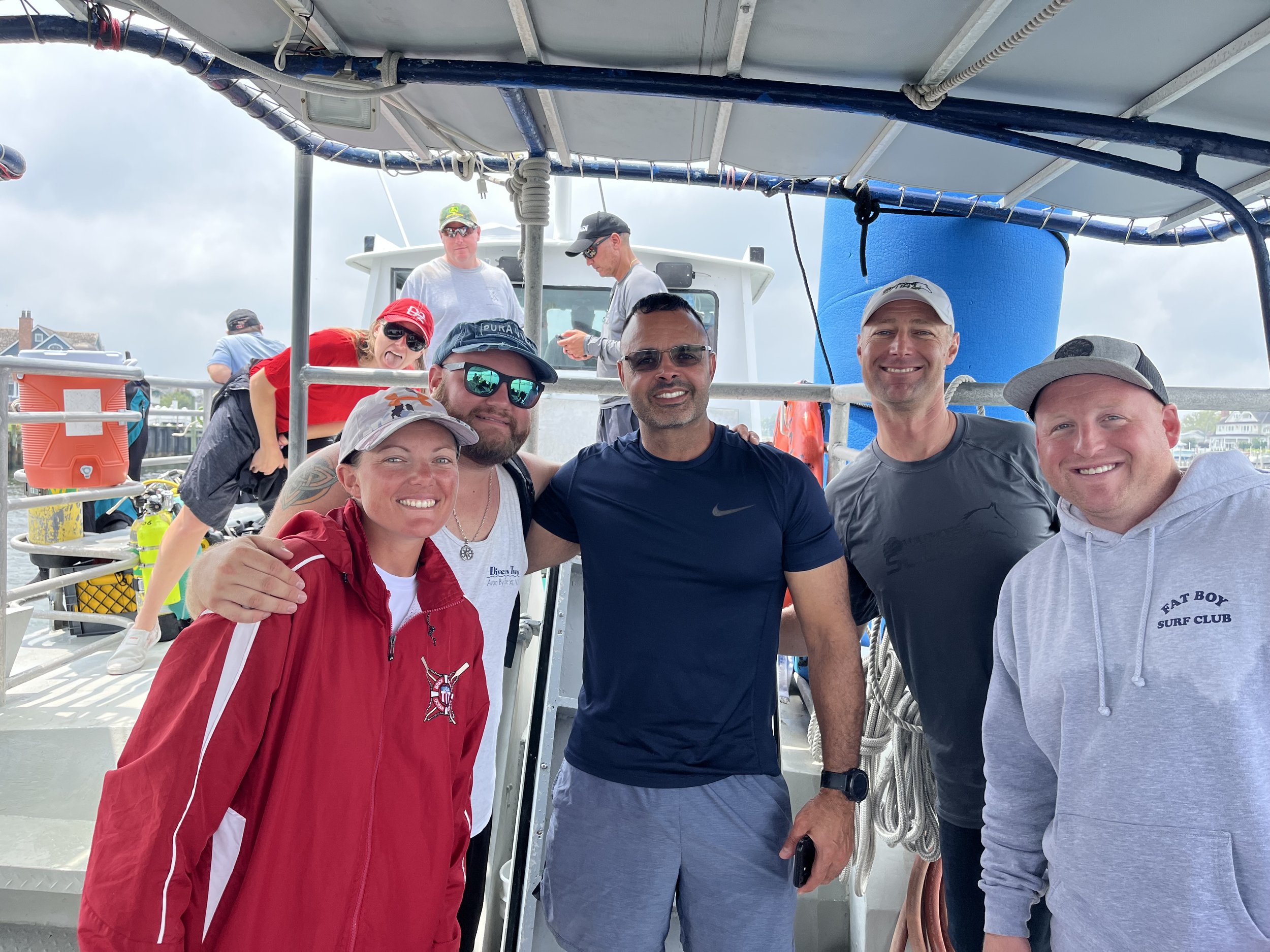 Group of six people smiling on a boat during a daytime outing. They are dressed casually in sportswear, with some wearing hats and sunglasses. The background includes the boat's structure and other individuals, with cloudy skies overhead.