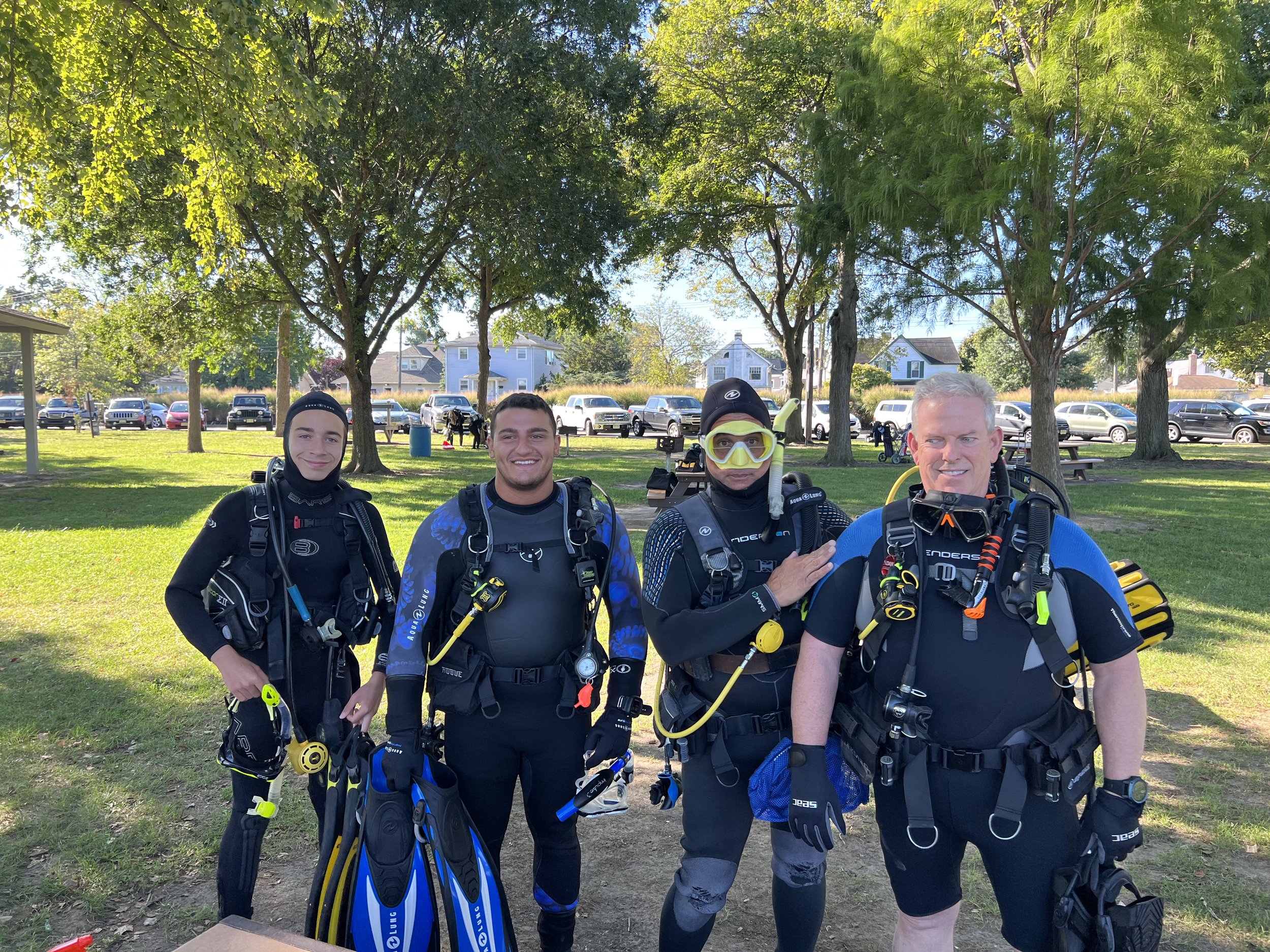 Group of four scuba divers wearing wetsuits and gear, standing outdoors in a park with trees and parked cars in the background.
