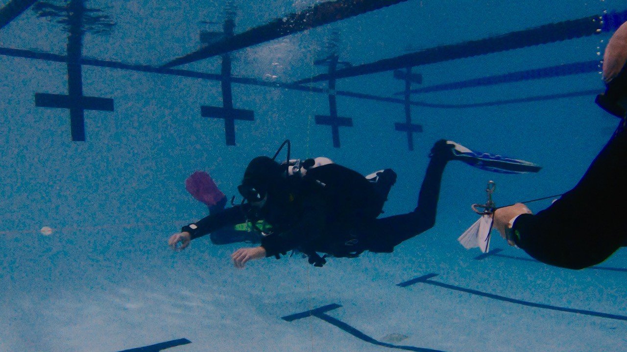 Person scuba diving underwater in a swimming pool, holding onto a line held by a person outside the pool. Depth markings are visible on the pool wall.