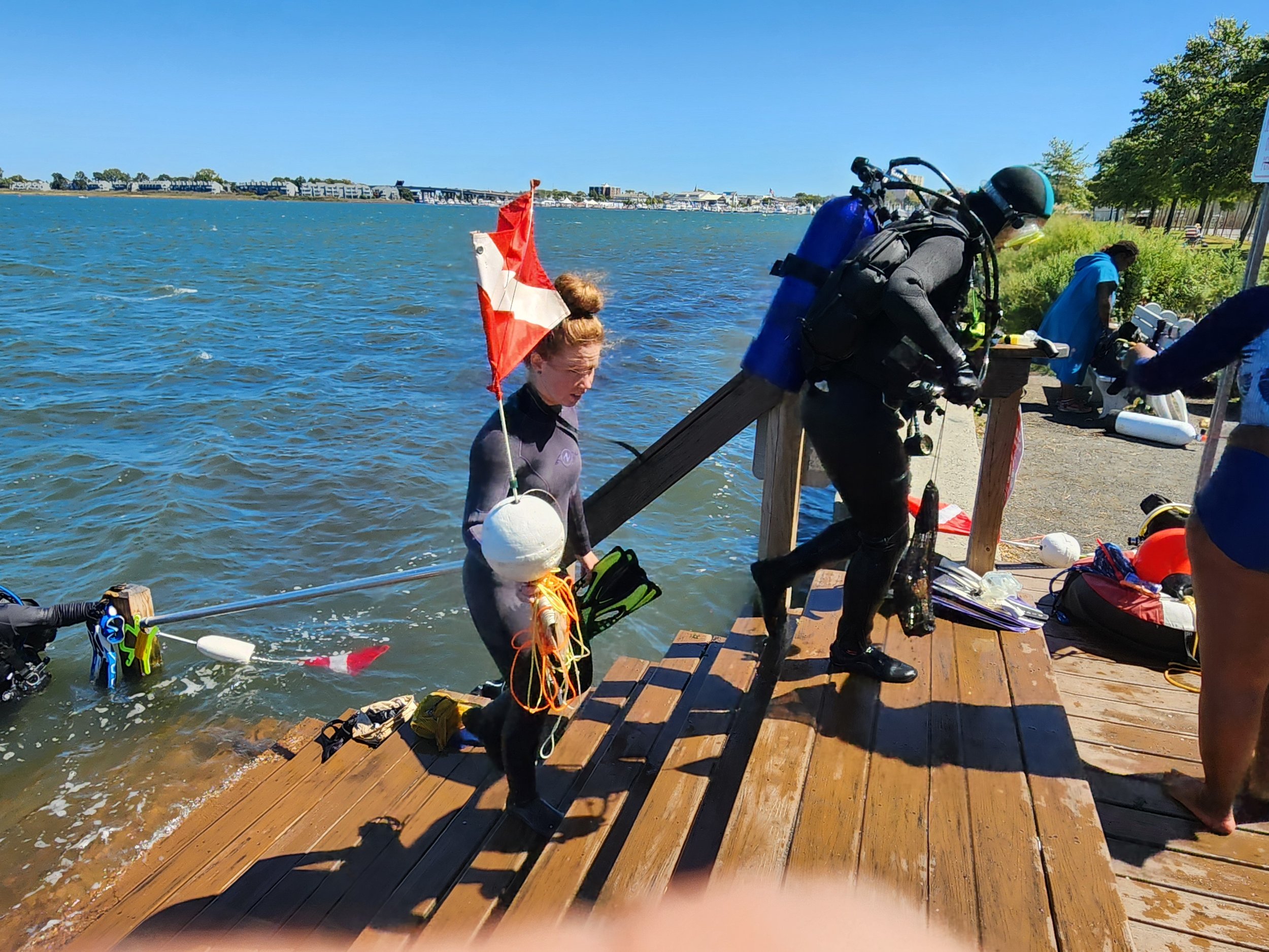 Scuba diver and young girl on a wooden dock preparing to enter a body of water; the girl holds a Canadian flag and floatation device, the diver is in a wetsuit with scuba gear, others are in the background near equipment.
