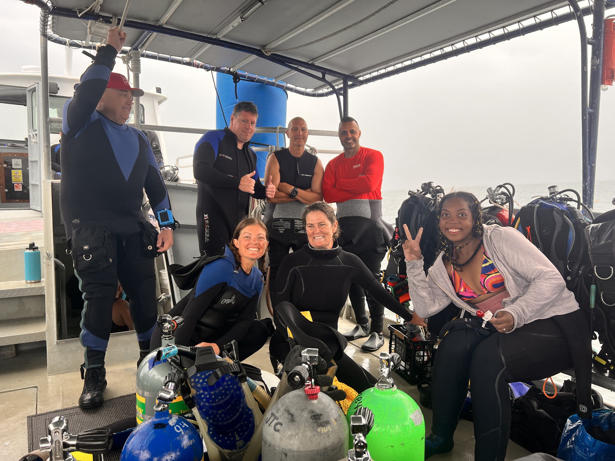 Group of eight people preparing for scuba diving on a boat, surrounded by scuba tanks and gear.