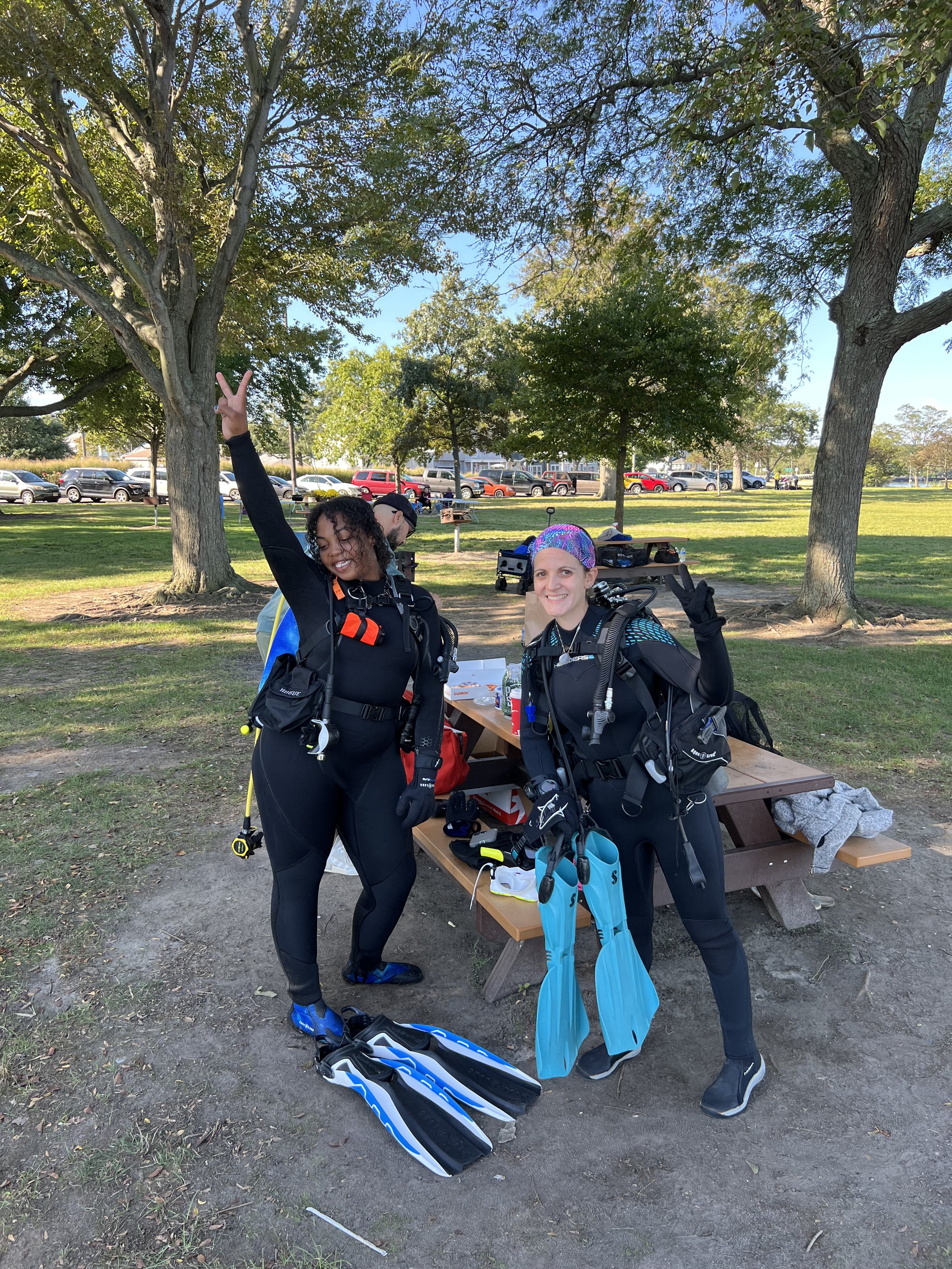 Two women in scuba diving gear smiling and posing with peace signs during an outdoor event in a park, with trees, cars, and a picnic table in the background.