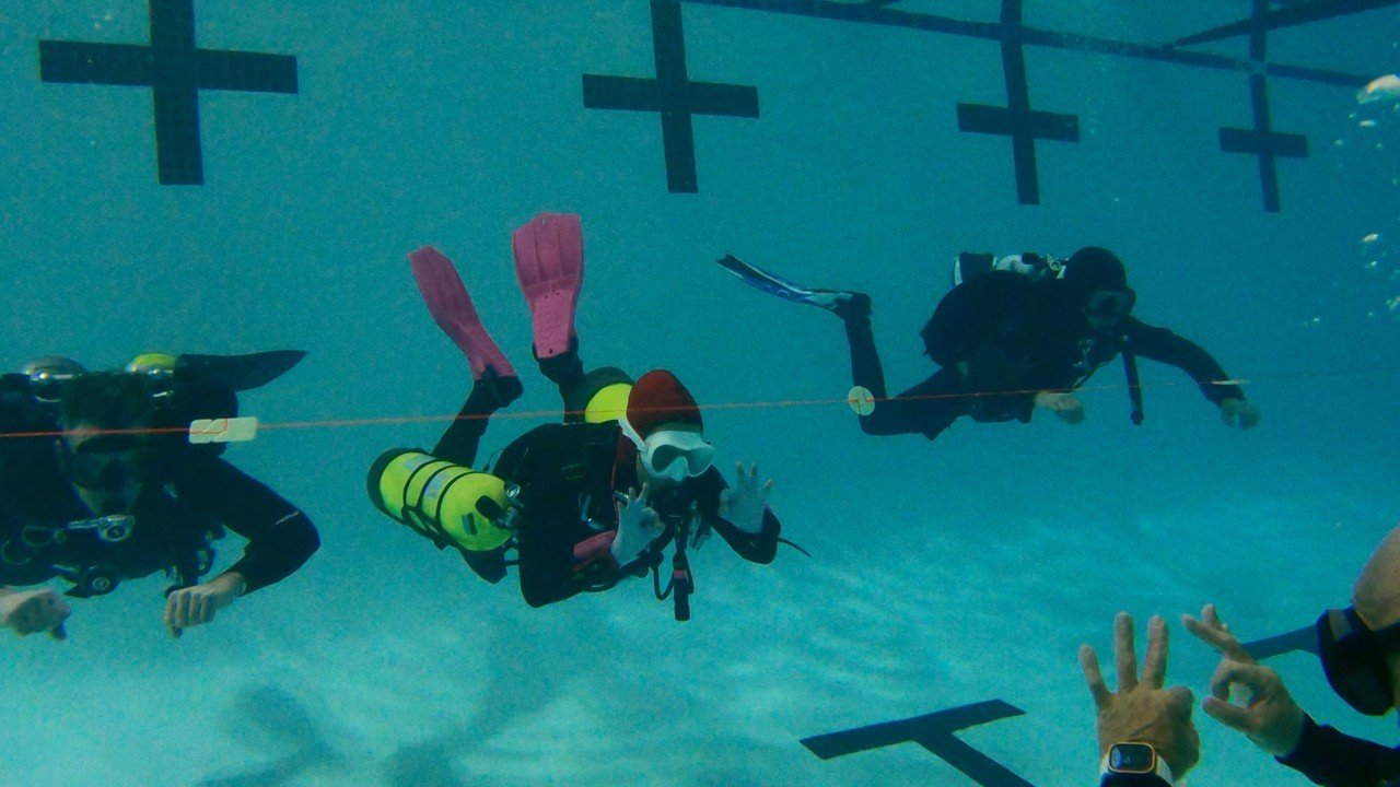 Three scuba divers underwater, one giving the peace sign, wearing masks and fins.