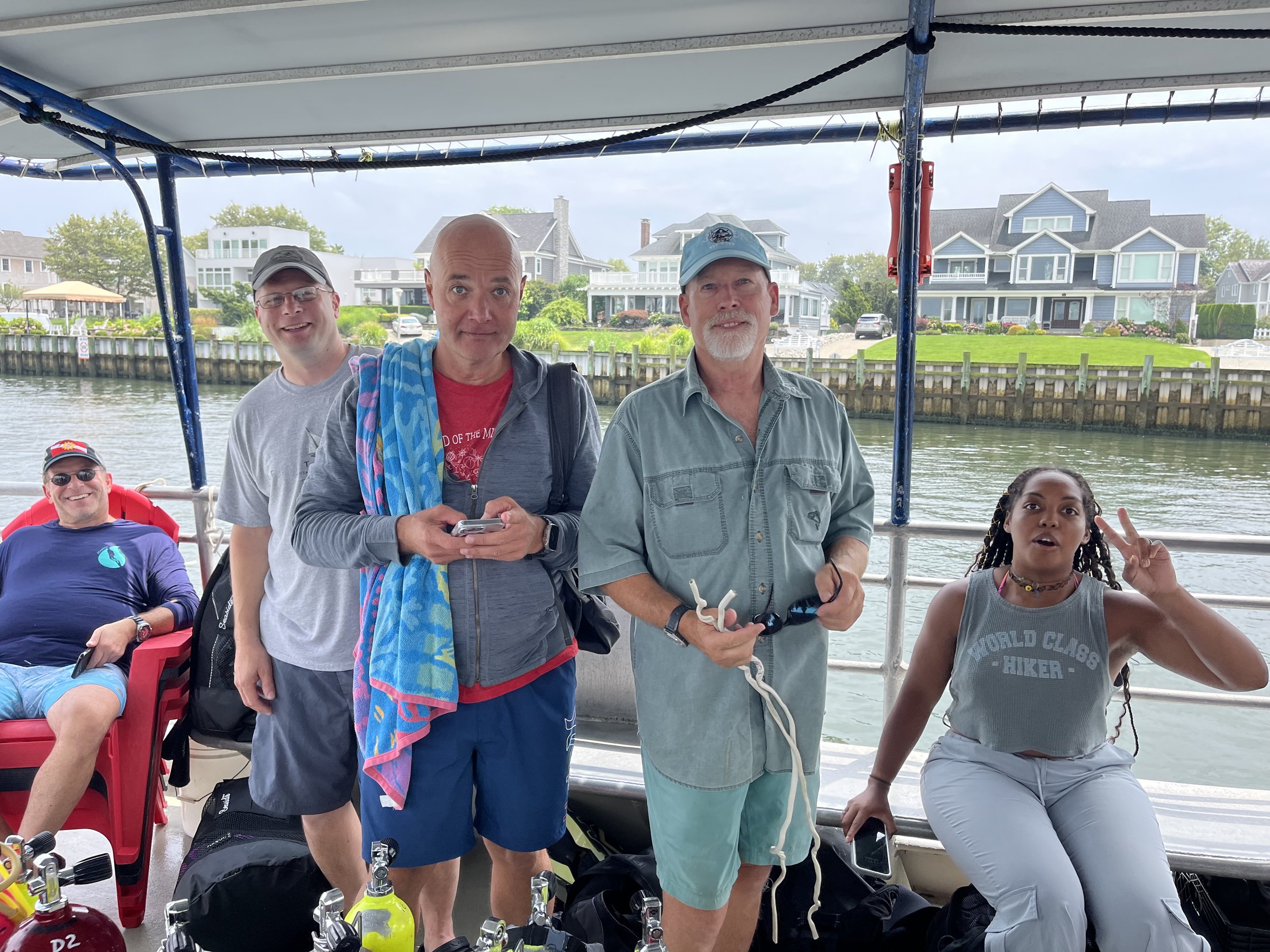 Group of five people on a boat, with water and houses in the background. They are dressed casually, some wearing sunglasses, with one person sitting and four standing. One person is making a peace sign. The setting appears to be a daytime boat trip.