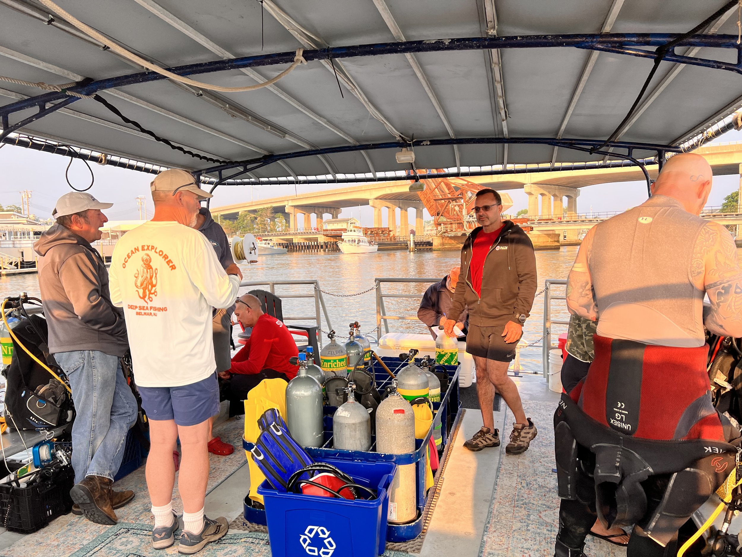 A group of people on a boat preparing for scuba diving or diving activity, with scuba tanks and gear, near a body of water and a bridge in the background.
